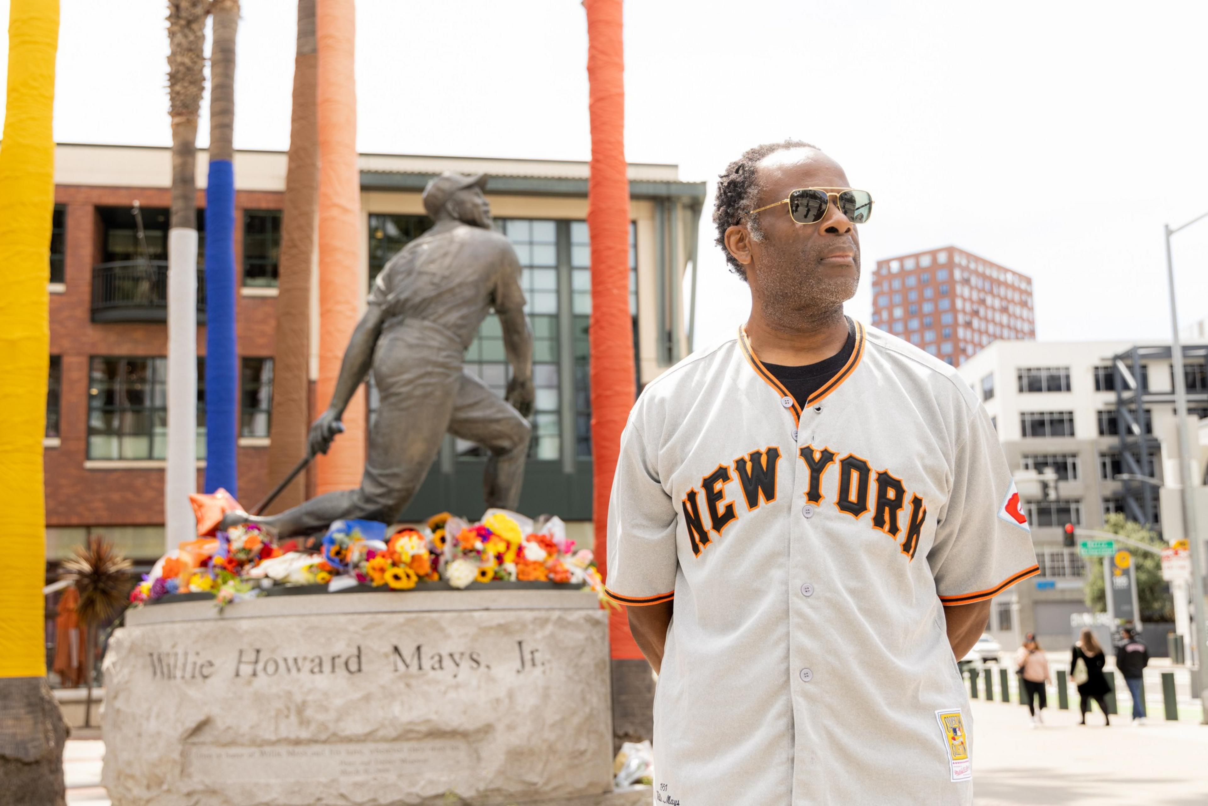 A man in a &quot;New York&quot; baseball jersey stands in front of a Willie Howard Mays Jr. statue adorned with colorful flowers. The backdrop includes modern buildings and wrapped palm trees.