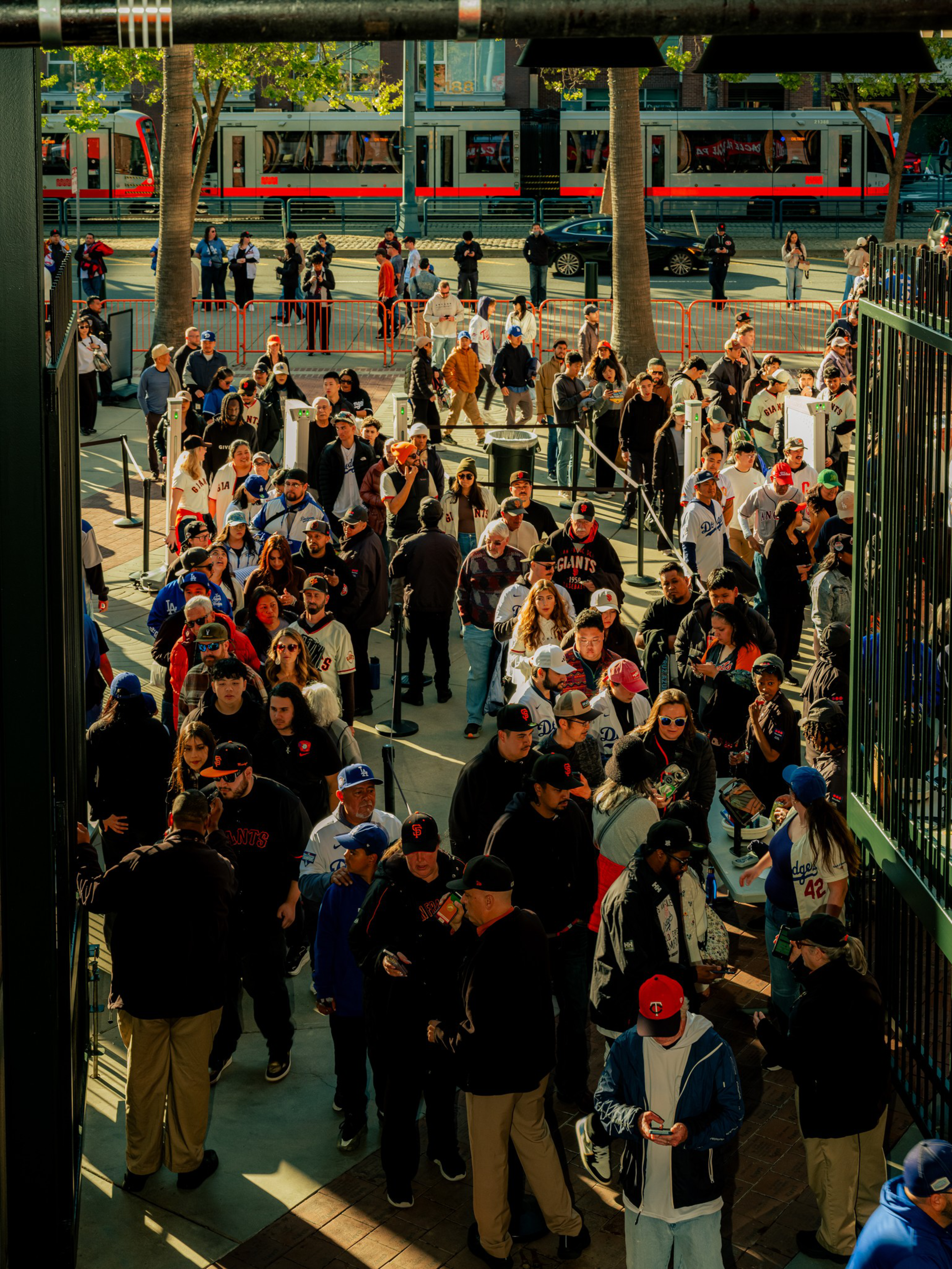 A large crowd of people, many wearing baseball jerseys and caps, waits in line outside a venue with trees and a passing train in the background.