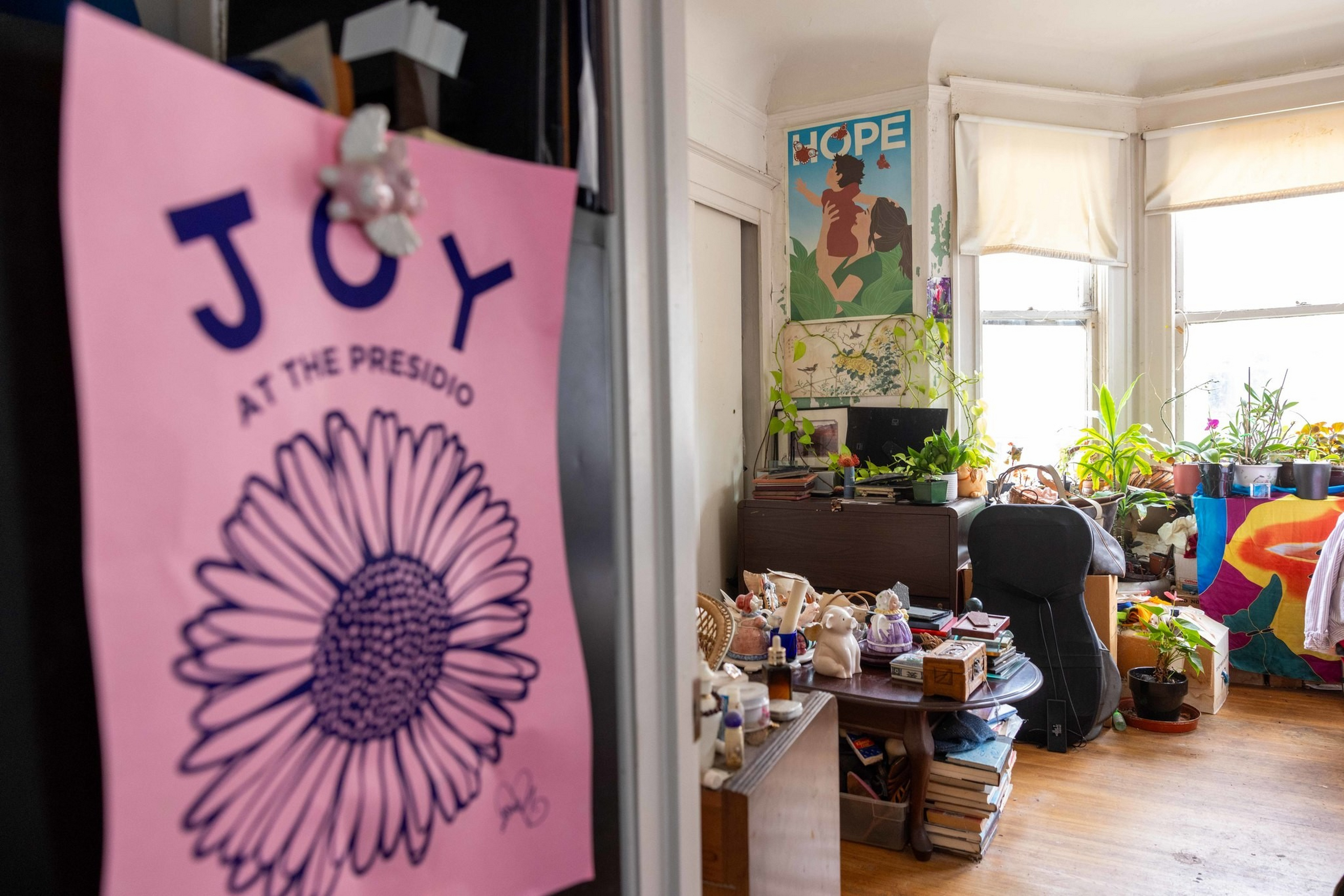 A cozy room filled with numerous potted plants, books stacked on shelves, a black chair, a colorful blanket, and posters on the walls.