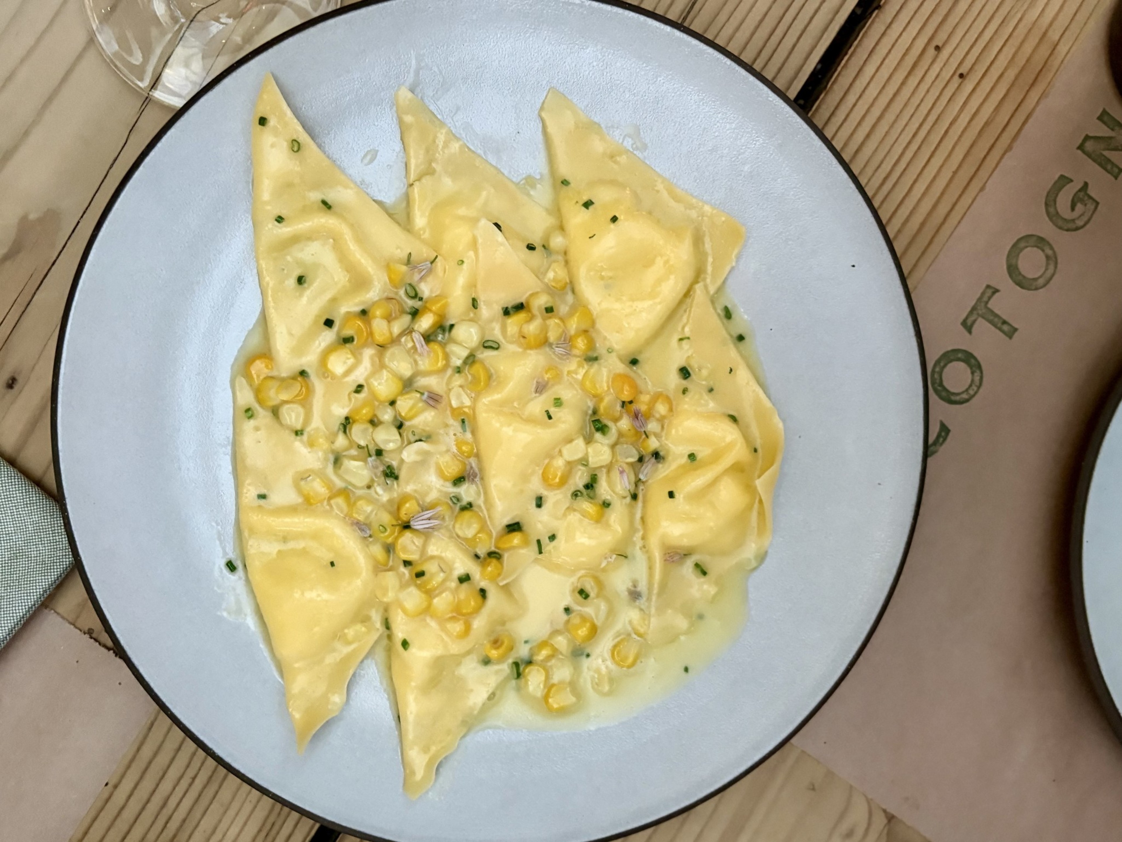 Four large triangular ravioli pieces are served on a white plate with a creamy sauce, corn kernels, chopped herbs, and edible flower petals.