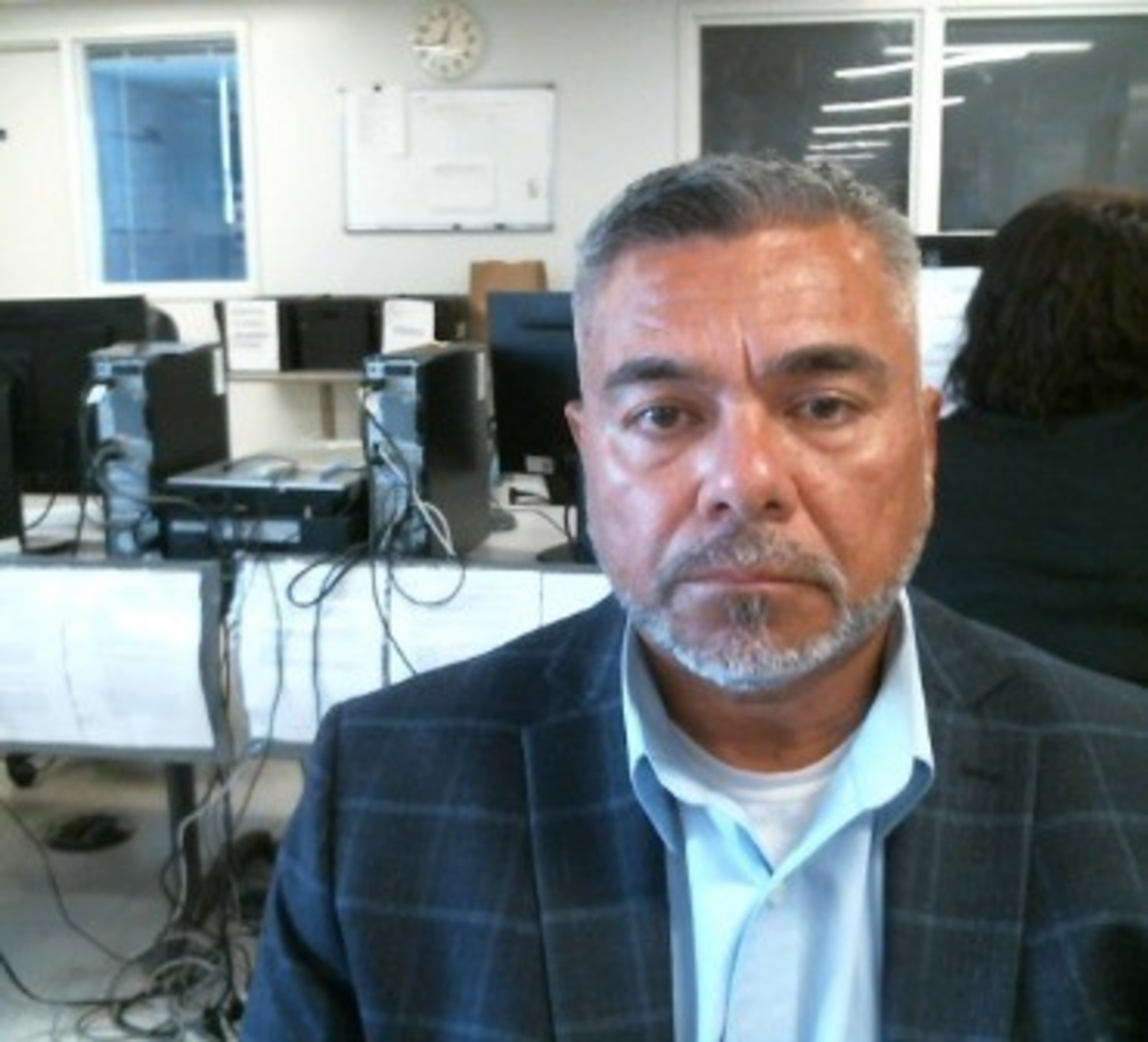 A man with gray hair and a trimmed beard wears a checkered blazer and light blue shirt, sitting in an office with computers and a clock on the wall.