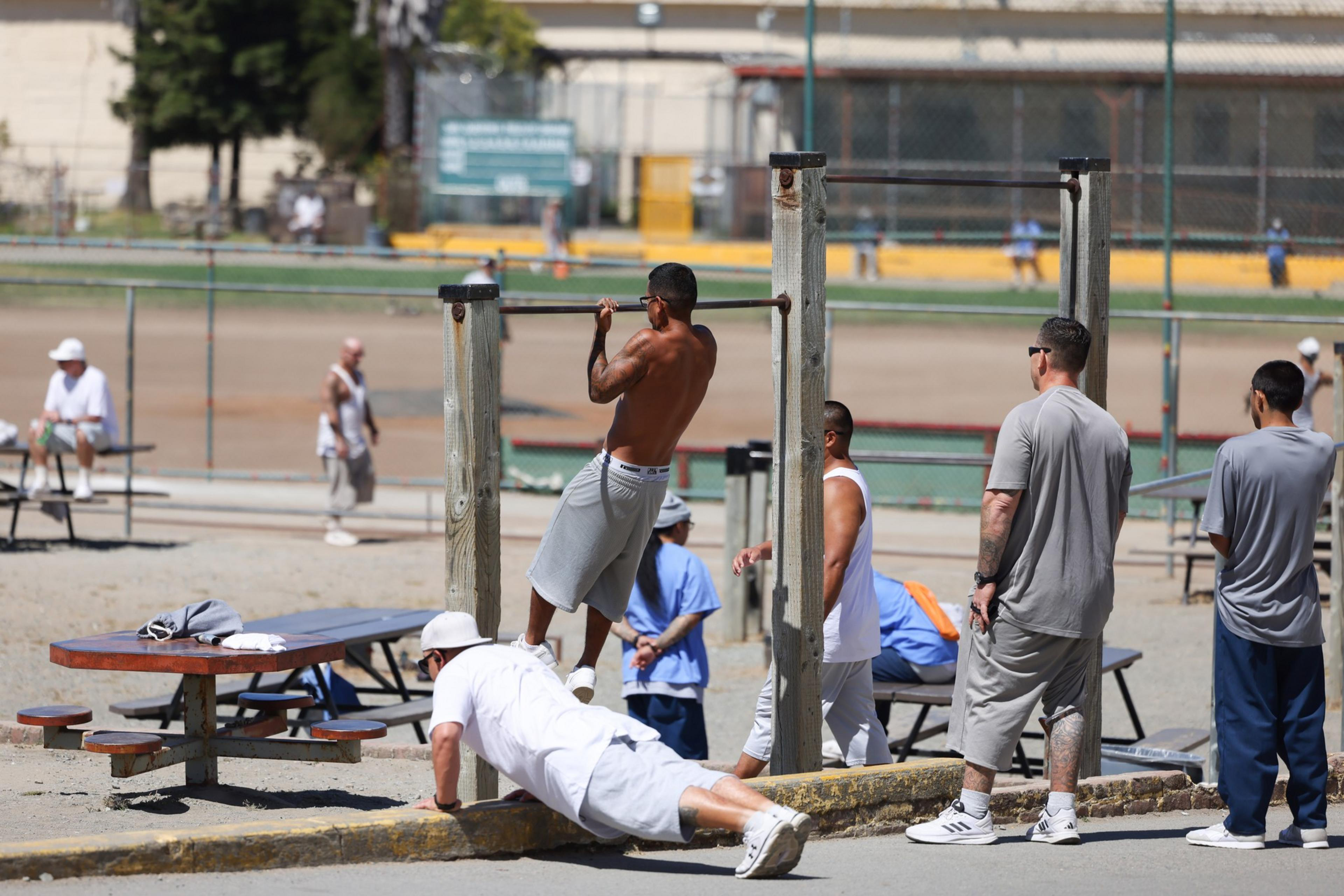 Men exercise at an outdoor gym; one does pull-ups while another does push-ups. Others watch or wait their turn.