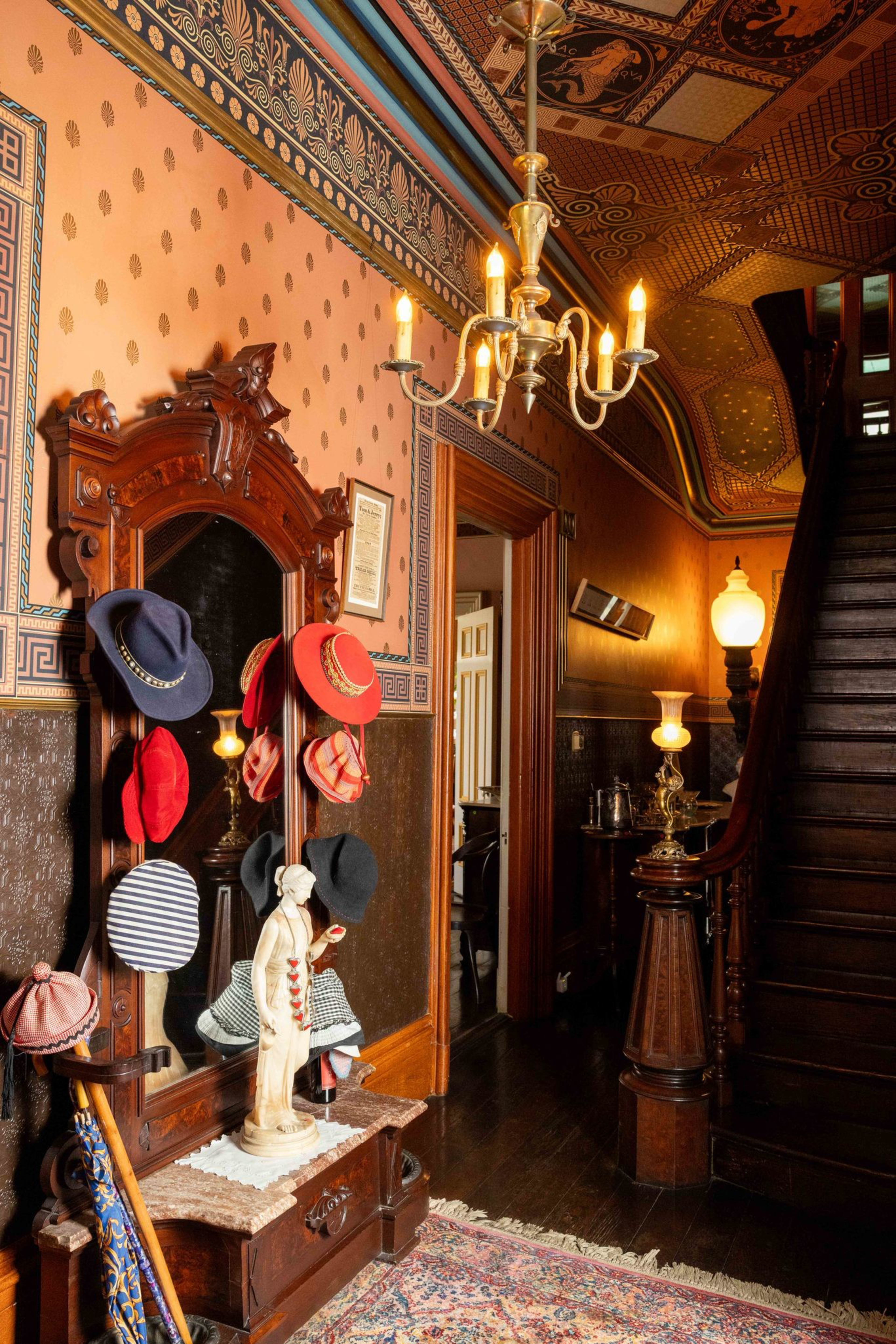 An ornate hallway features a wooden hat rack with various hats, a statue, a chandelier, a patterned rug, vintage lamps, and a wooden staircase.