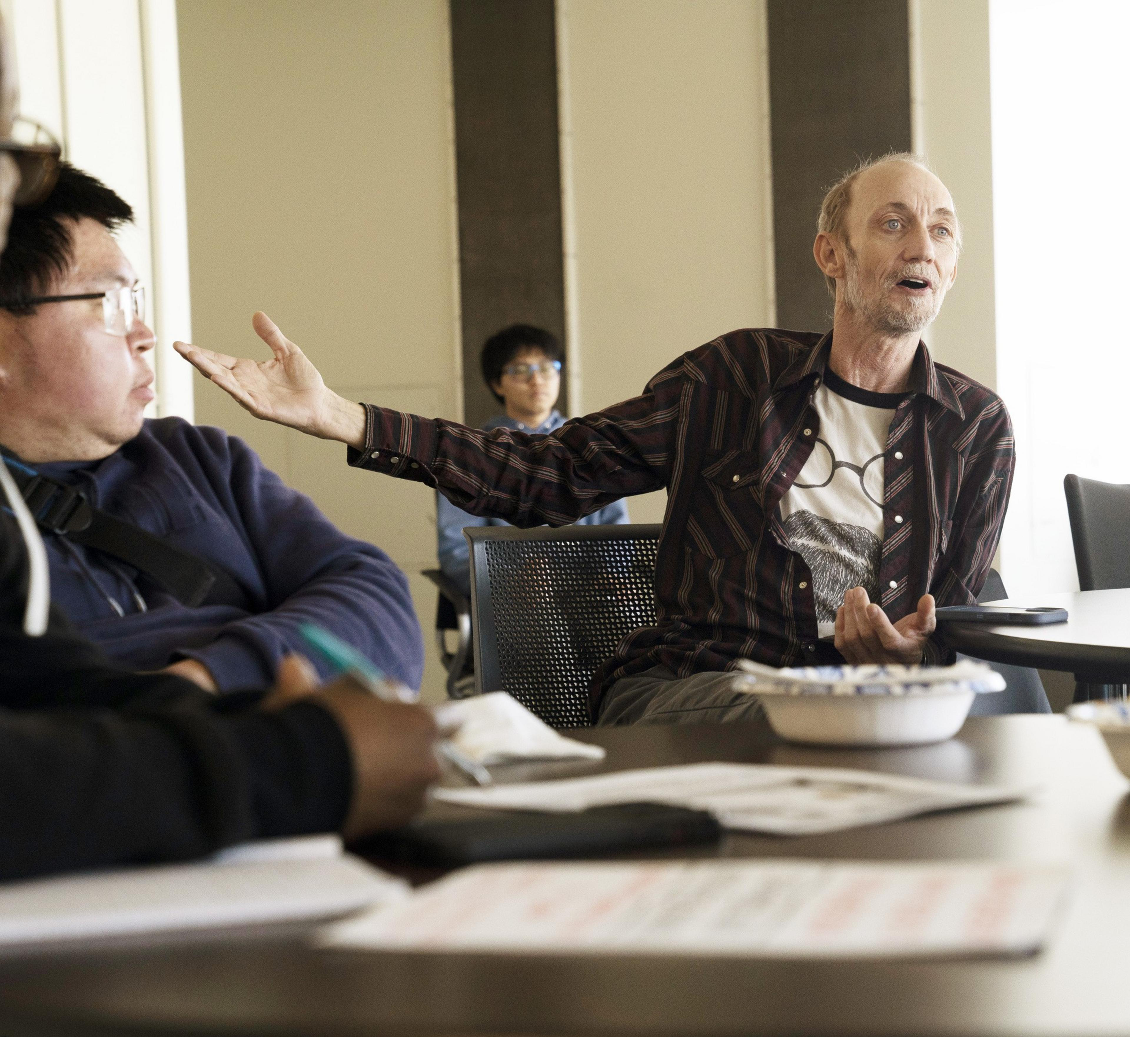 A man gestures passionately during a discussion in a classroom setting, engaging with another man seated nearby. Other people look on and listen attentively.