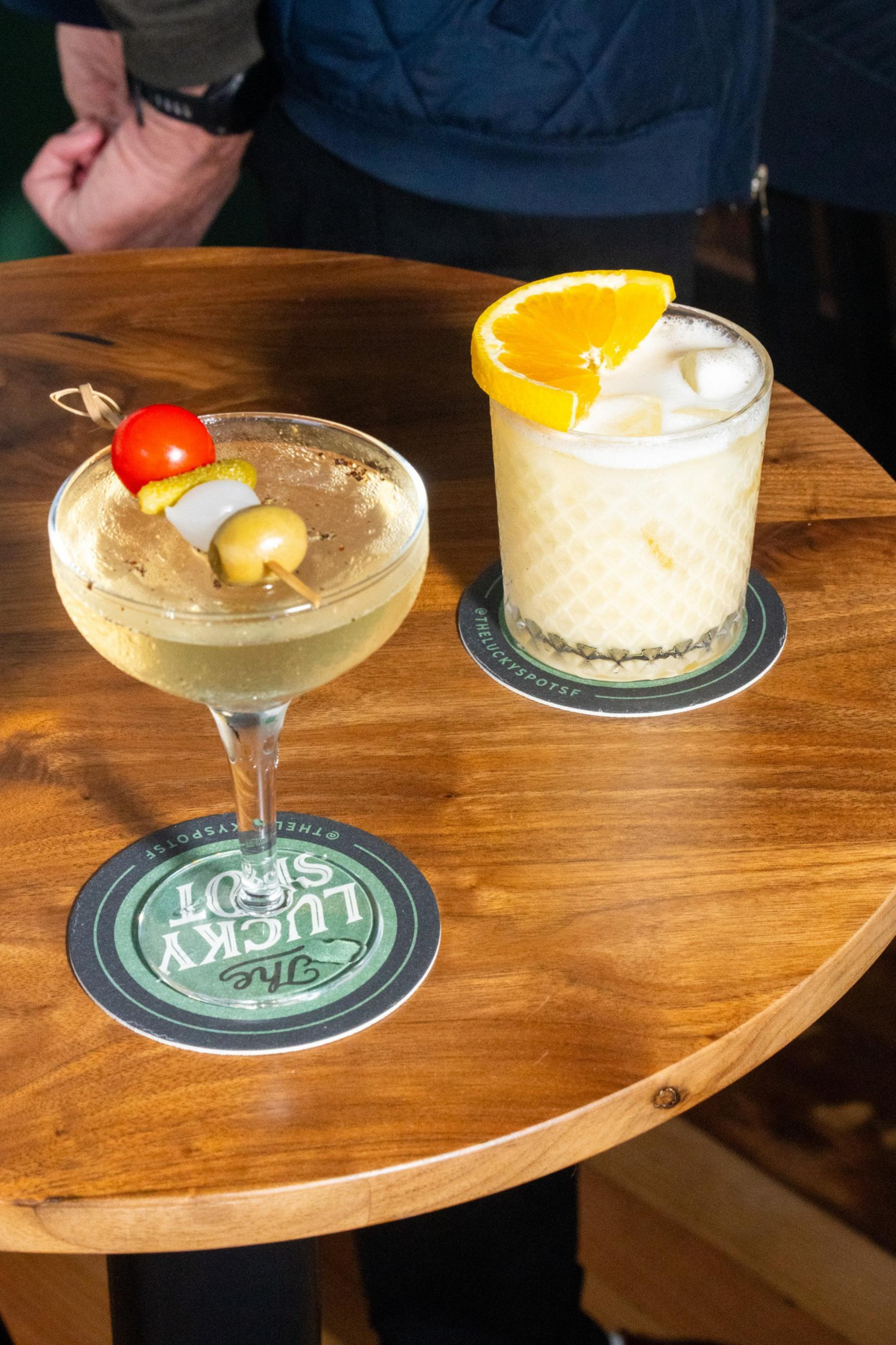 A clear cocktail with an olive, onion, pickle, and cherry tomato skewer sits next to a creamy drink garnished with an orange slice on a wooden table.