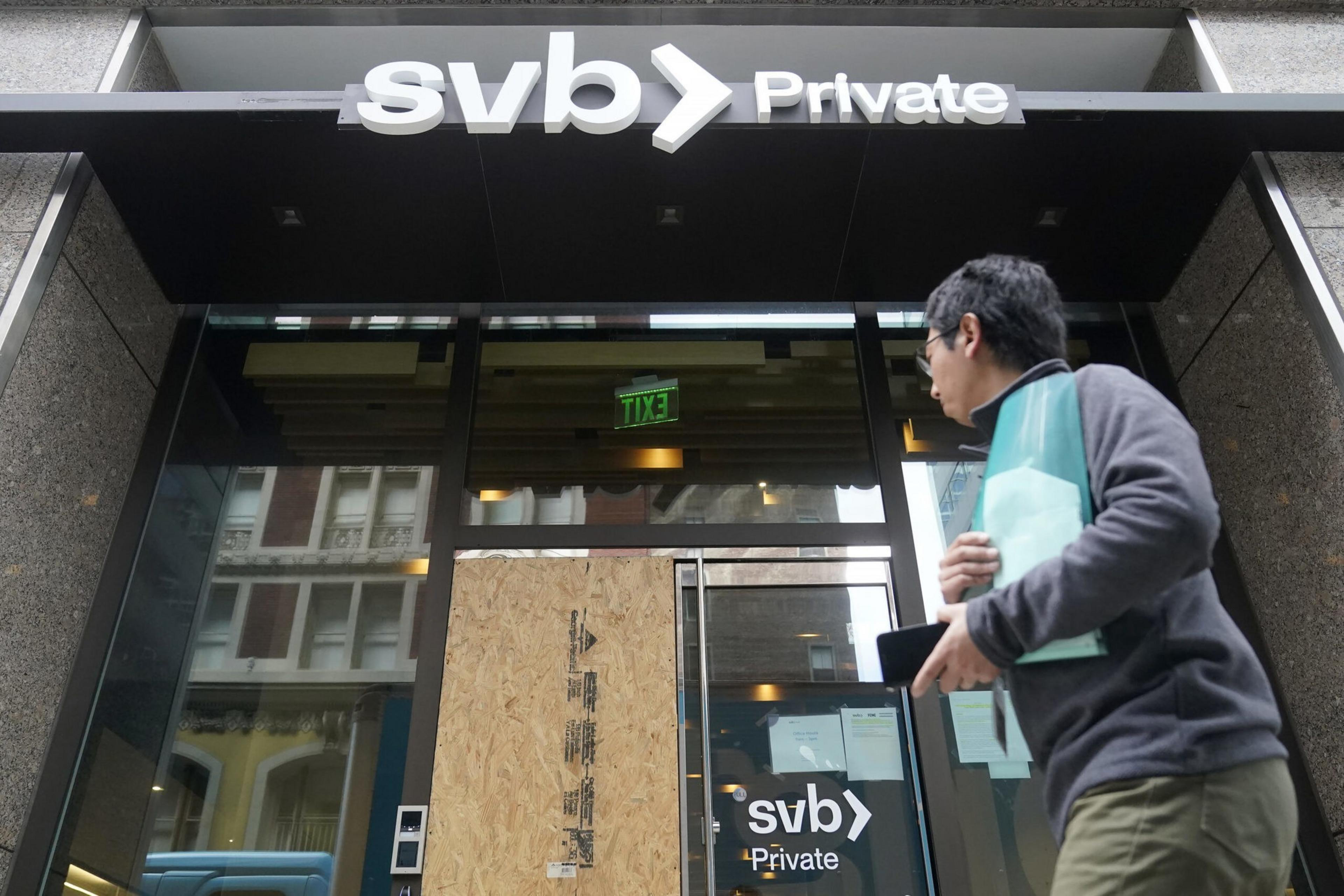 A man holding documents and a phone walks past a glass entrance with one door boarded up, under a large "svb Private" sign.