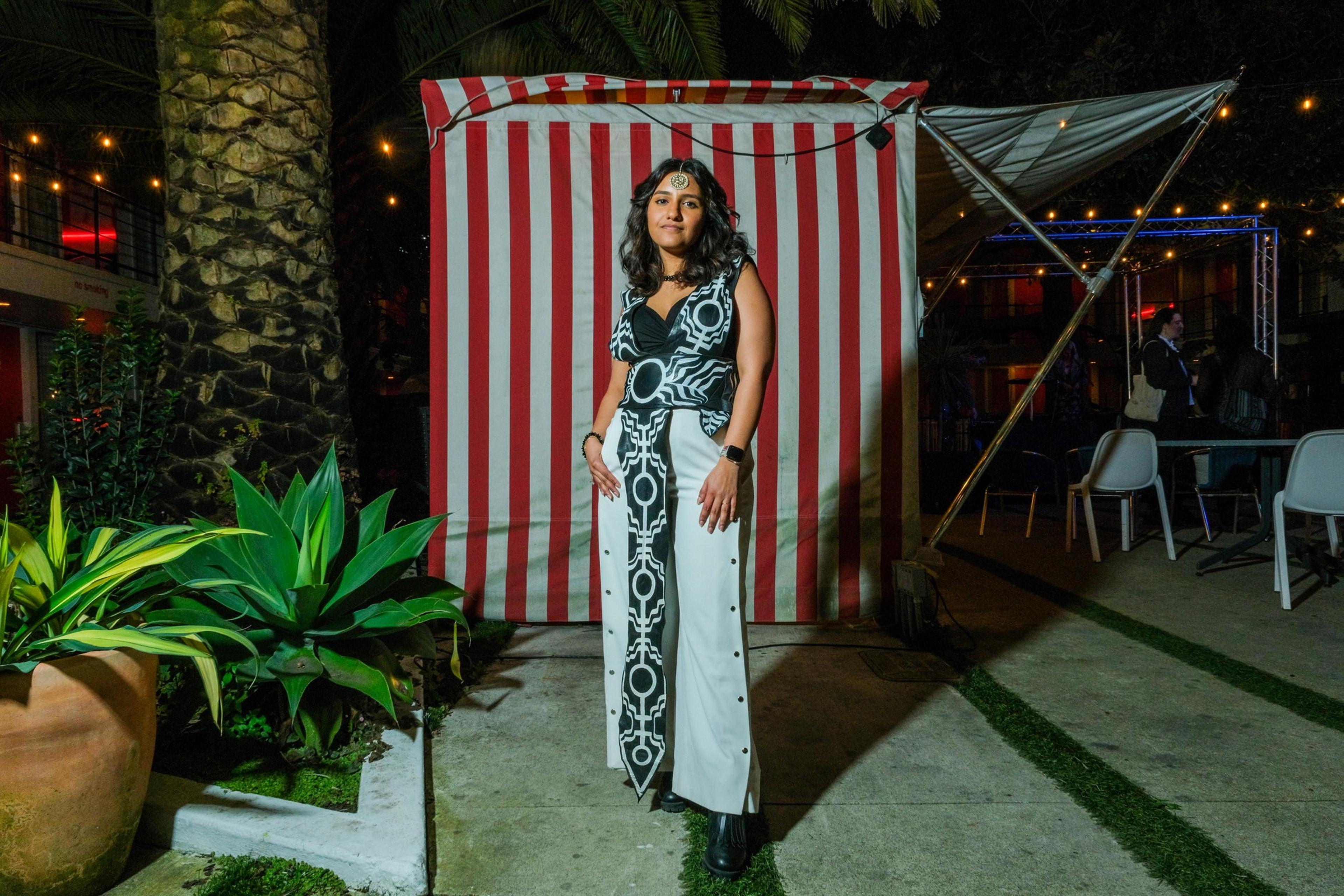 A person stands before a red &amp; white striped backdrop at night, wearing a patterned outfit and headpiece, with a party setting behind.