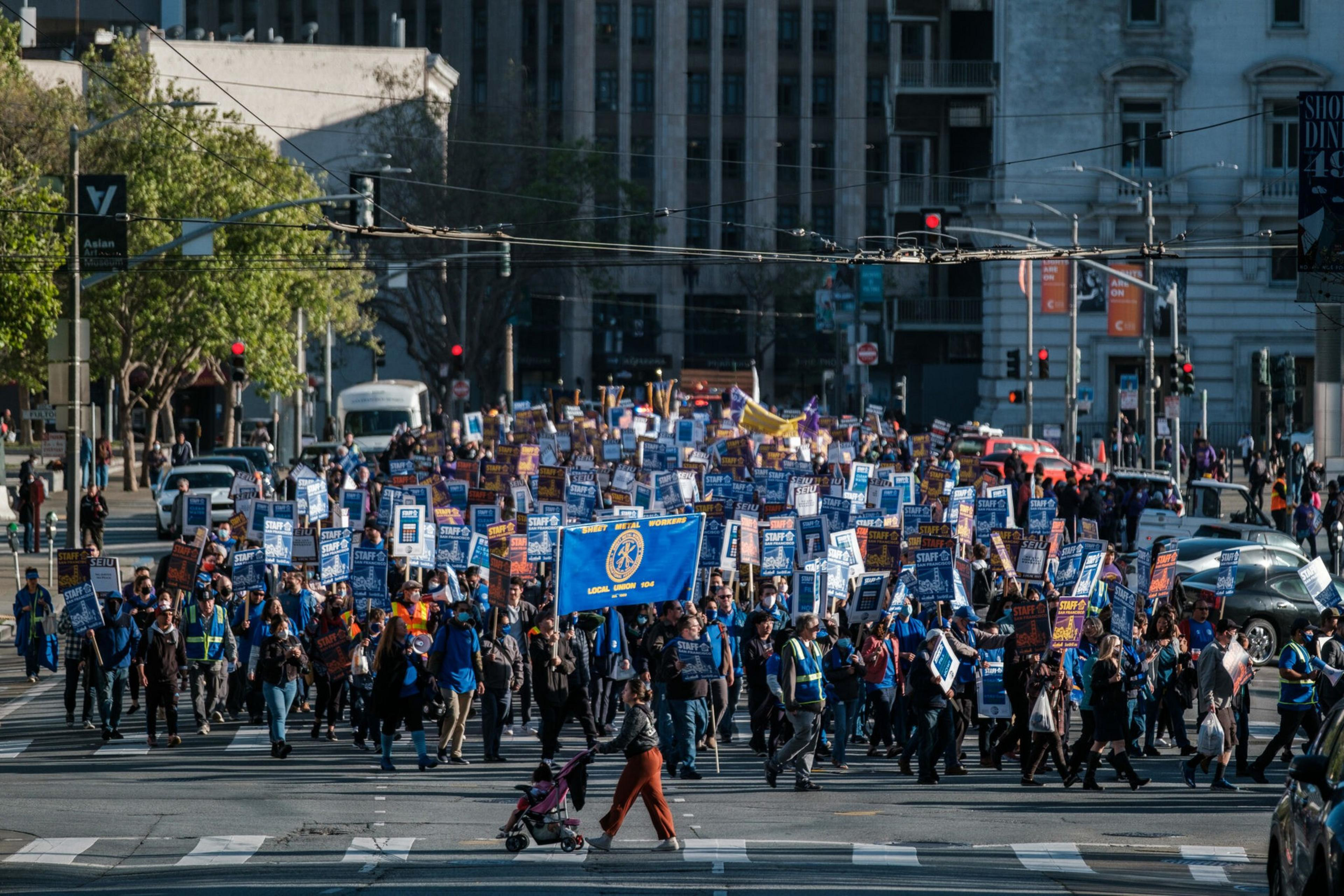 A large crowd of people marches through a city street holding signs and banners demanding a higher minimum wage as traffic lights turn red.