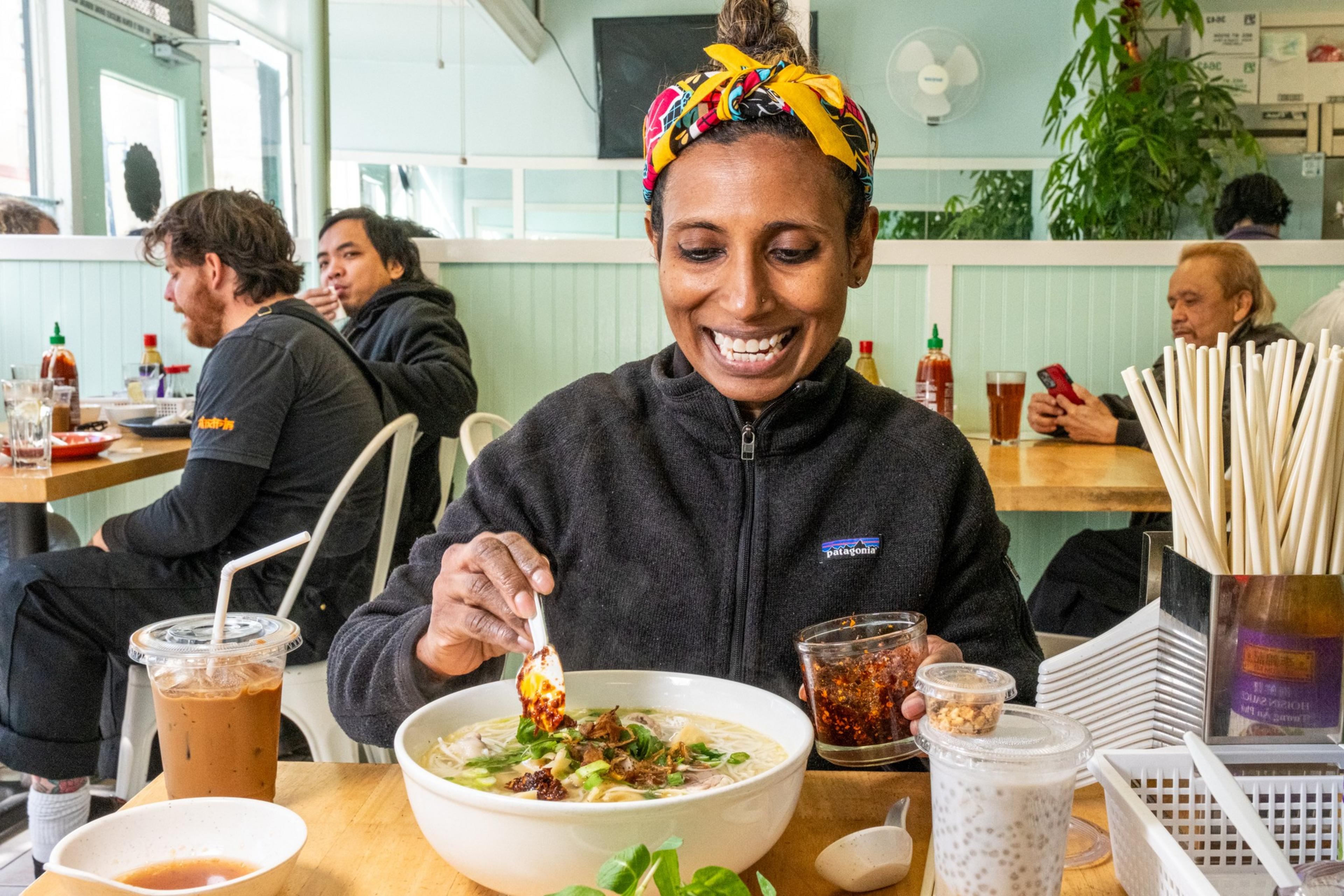 A woman with a vibrant headscarf smiles while adding chili to her noodle bowl at a restaurant, surrounded by drinks and diners at nearby tables.