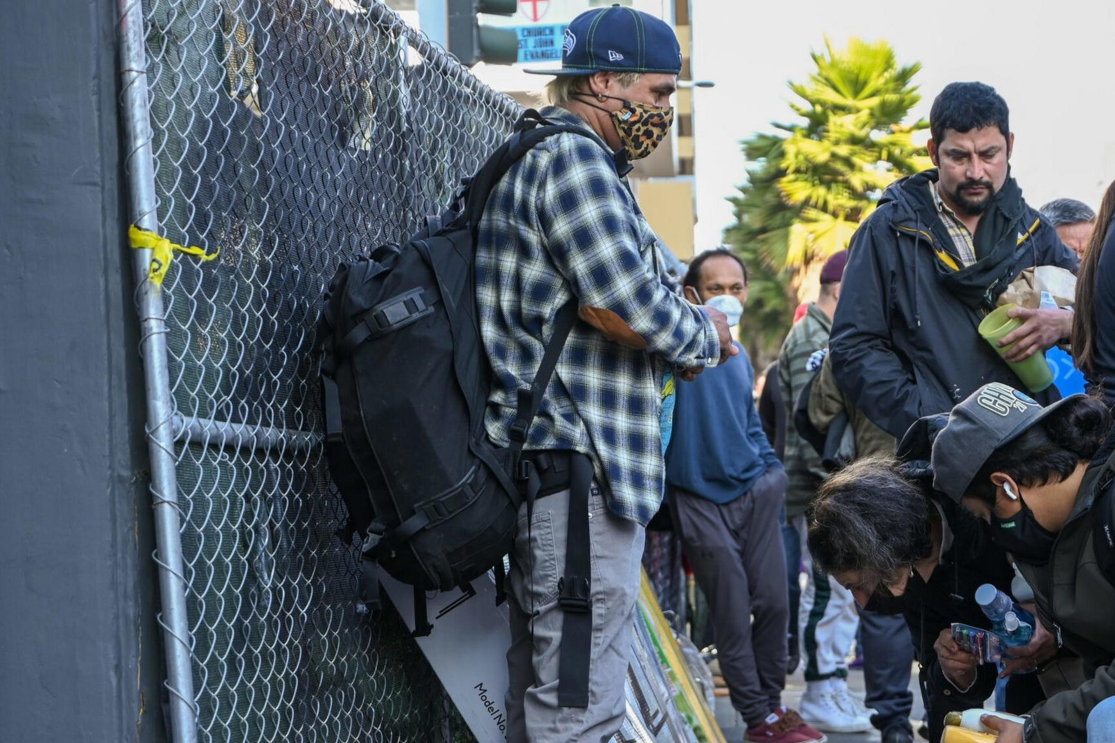 People, some wearing masks, are gathered on a sidewalk. One person crouches near an open suitcase. Others are holding items like water bottles and food.