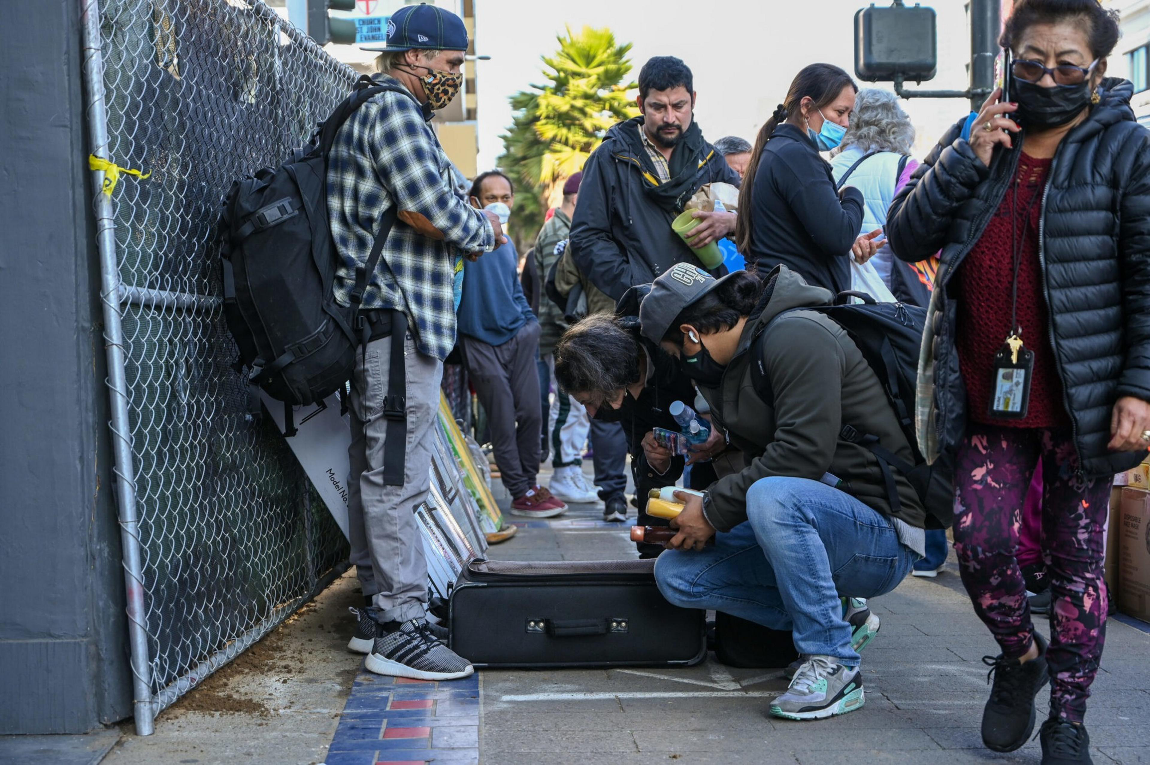 People, some wearing masks, are gathered on a sidewalk. One person crouches near an open suitcase. Others are holding items like water bottles and food.