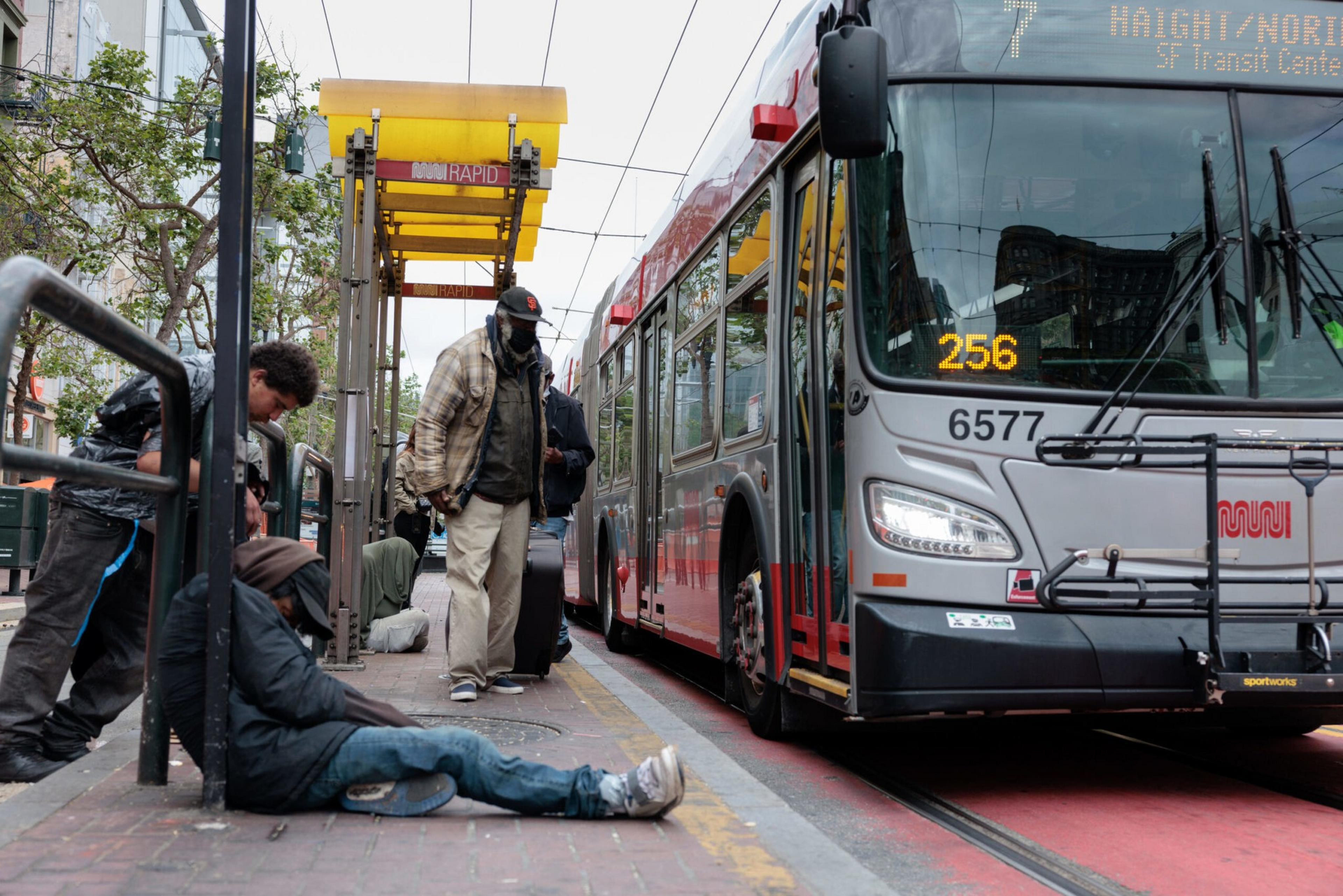 A bus labeled 256 is stopped at a Muni Rapid transit stop where several people, including one sitting on the ground, are waiting or walking.