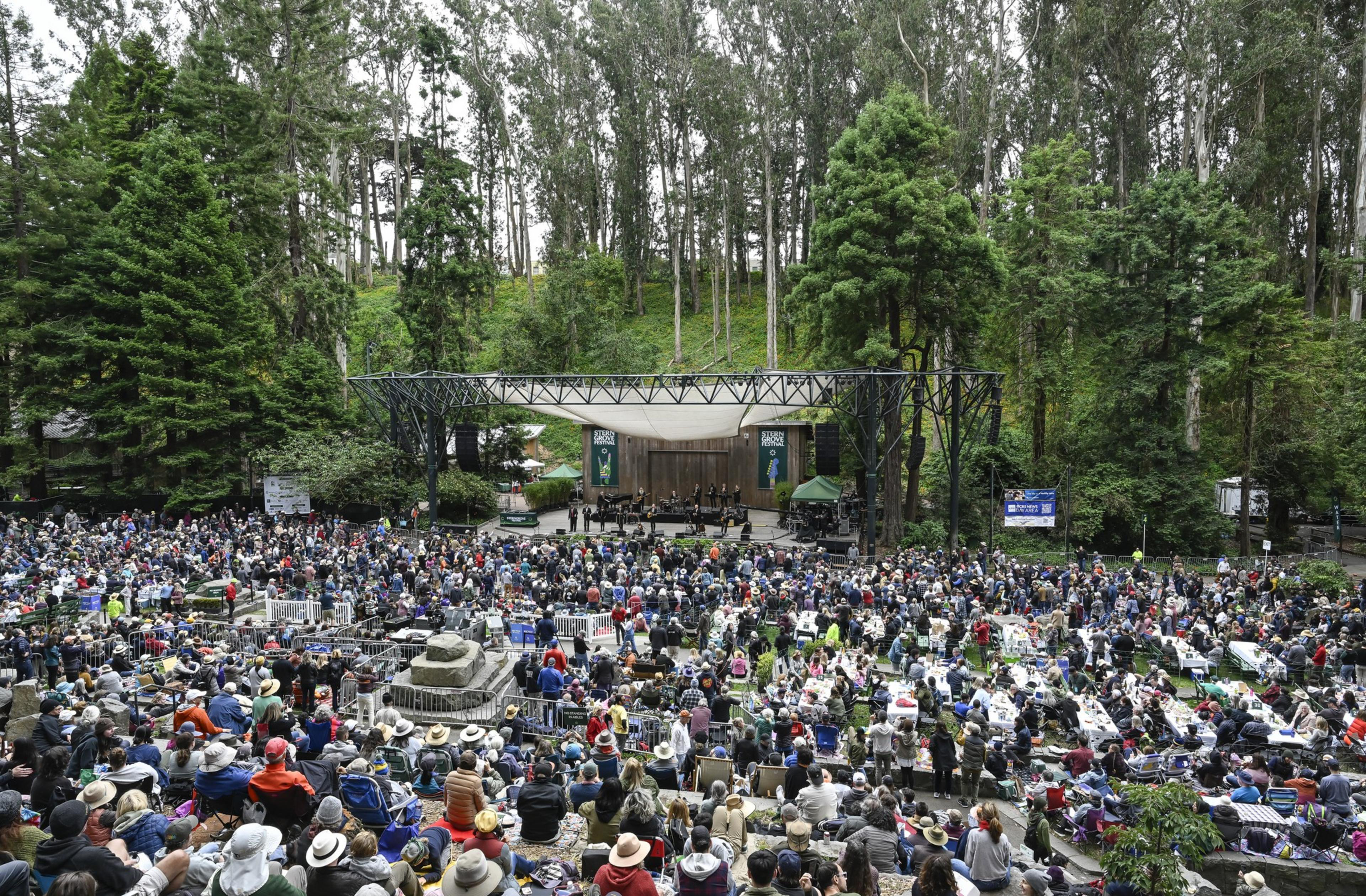 A large crowd gathers at an outdoor concert set in a forested area. People are seated on grass and stone benches, facing a stage with musicians performing.