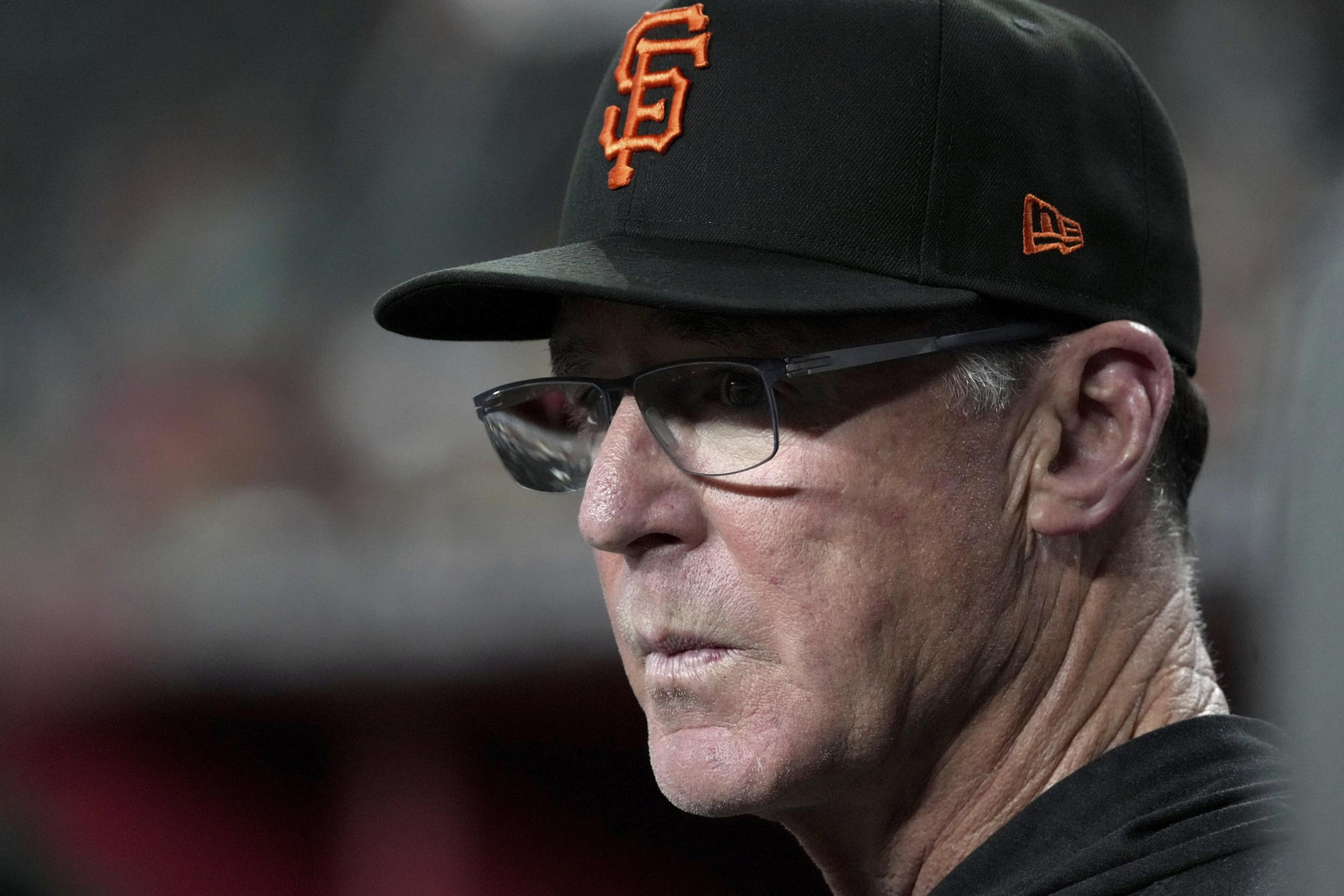 An older man wears glasses and a black baseball cap with an orange &quot;SF&quot; logo. His expression is focused, with a blurred background behind him.