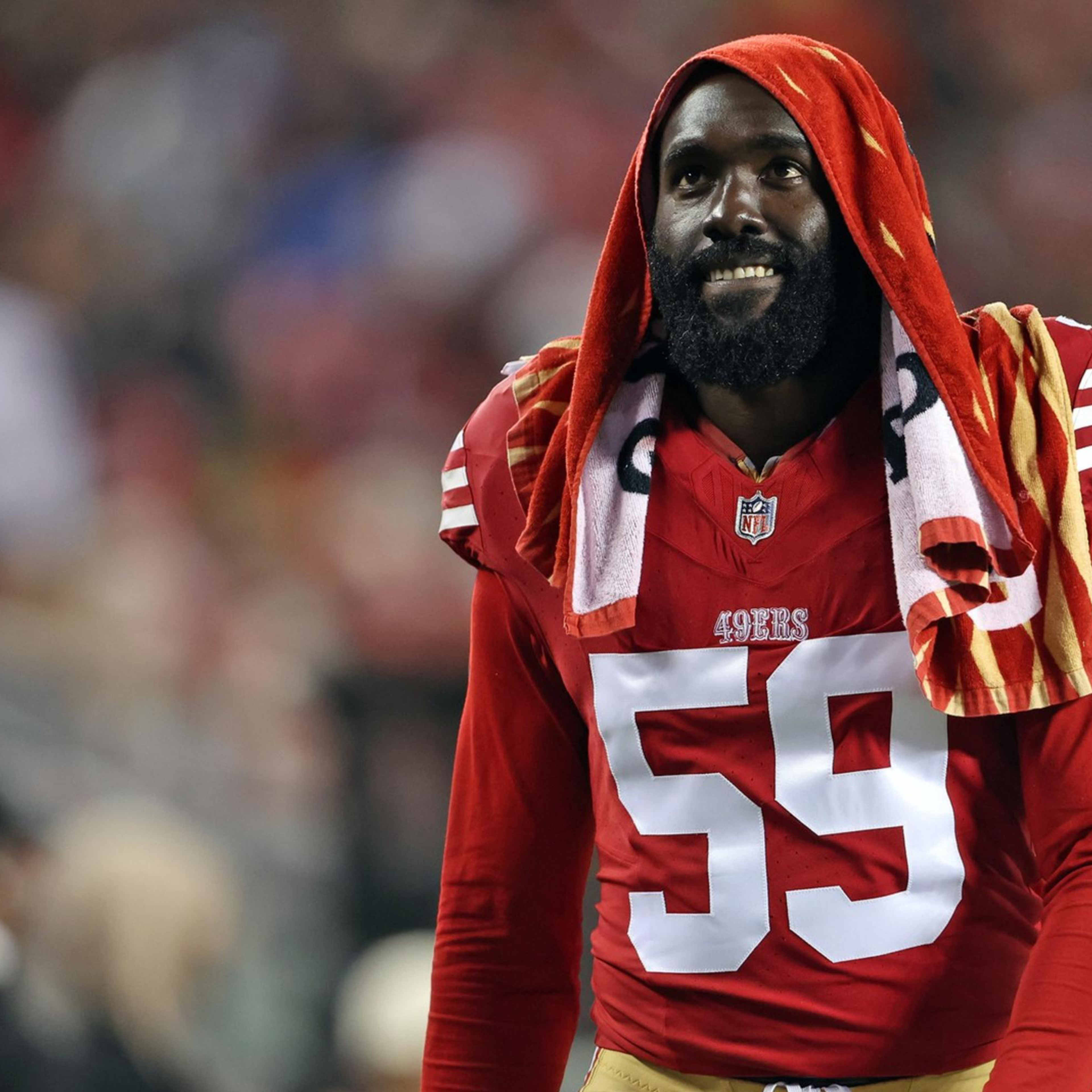 A football player wearing a red jersey with the number 59 stands on the sideline, a striped towel draped over his head. He is on a blurred field background.