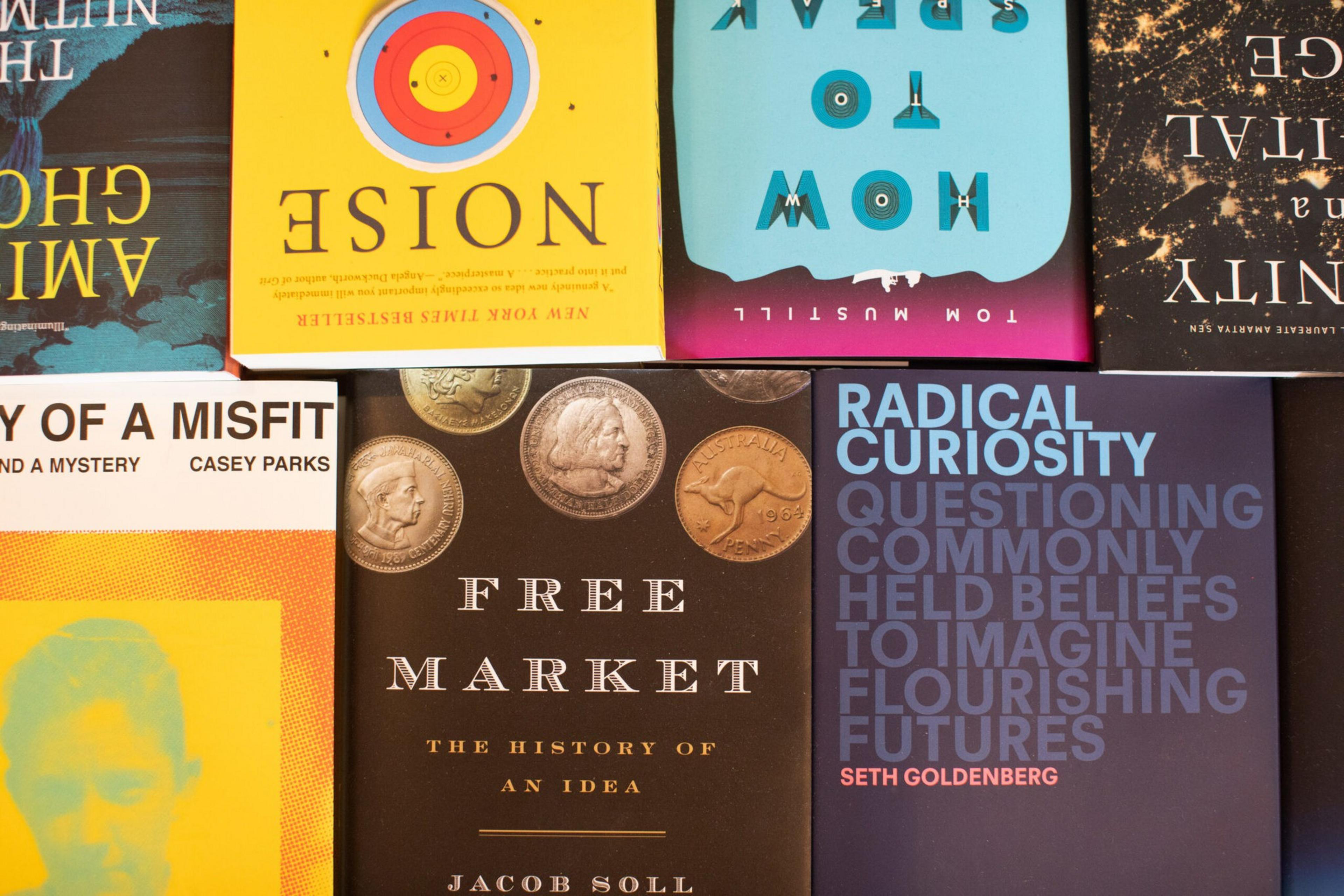 Several books are arranged, including titles like "Noise," "How to Speak," "Free Market," "Radical Curiosity," and "The Story of a Misfit," with coins featured on one cover.