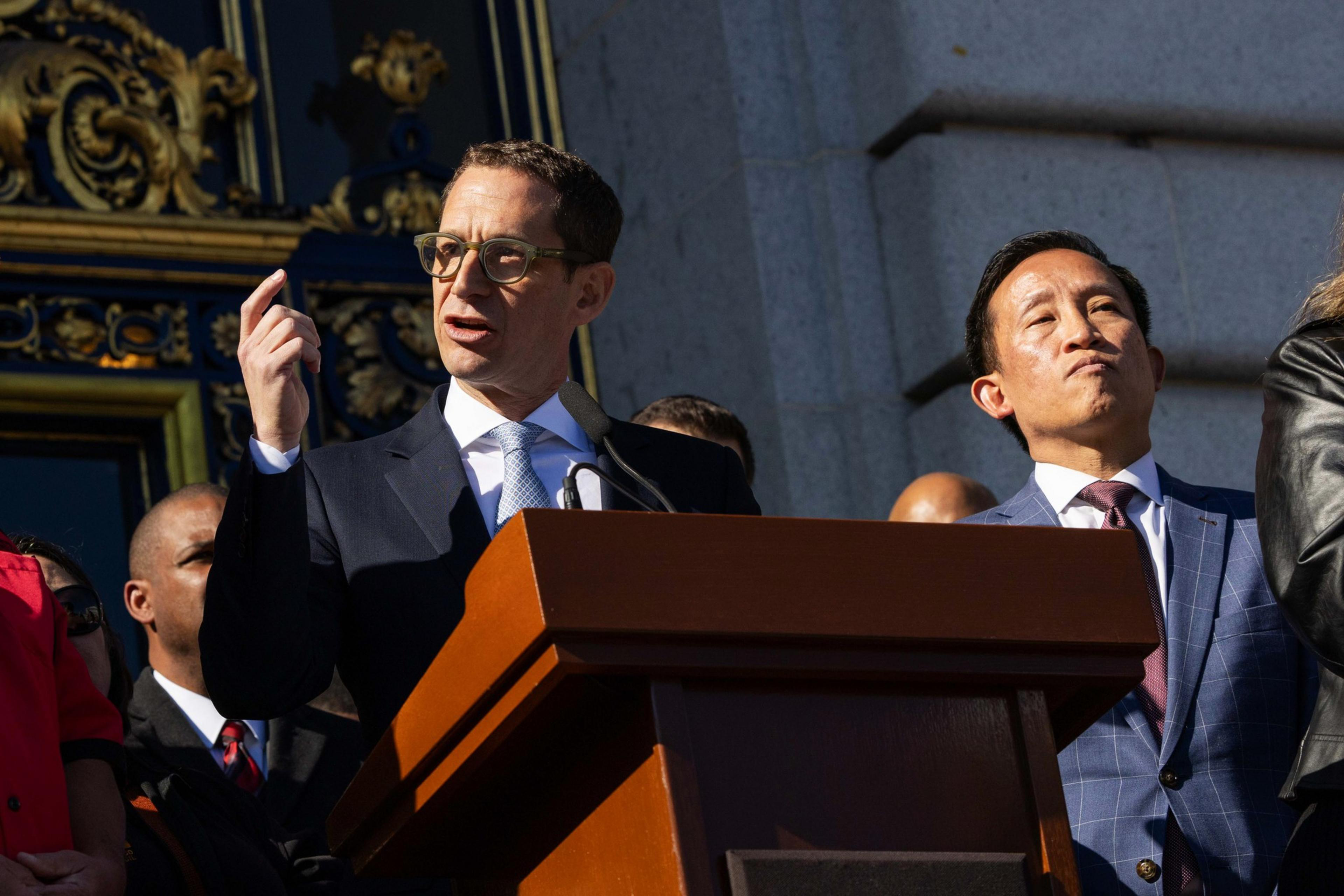 A man speaks passionately at a podium outdoors, wearing glasses and a suit. A second man stands beside him, listening intently in a formal setting.
