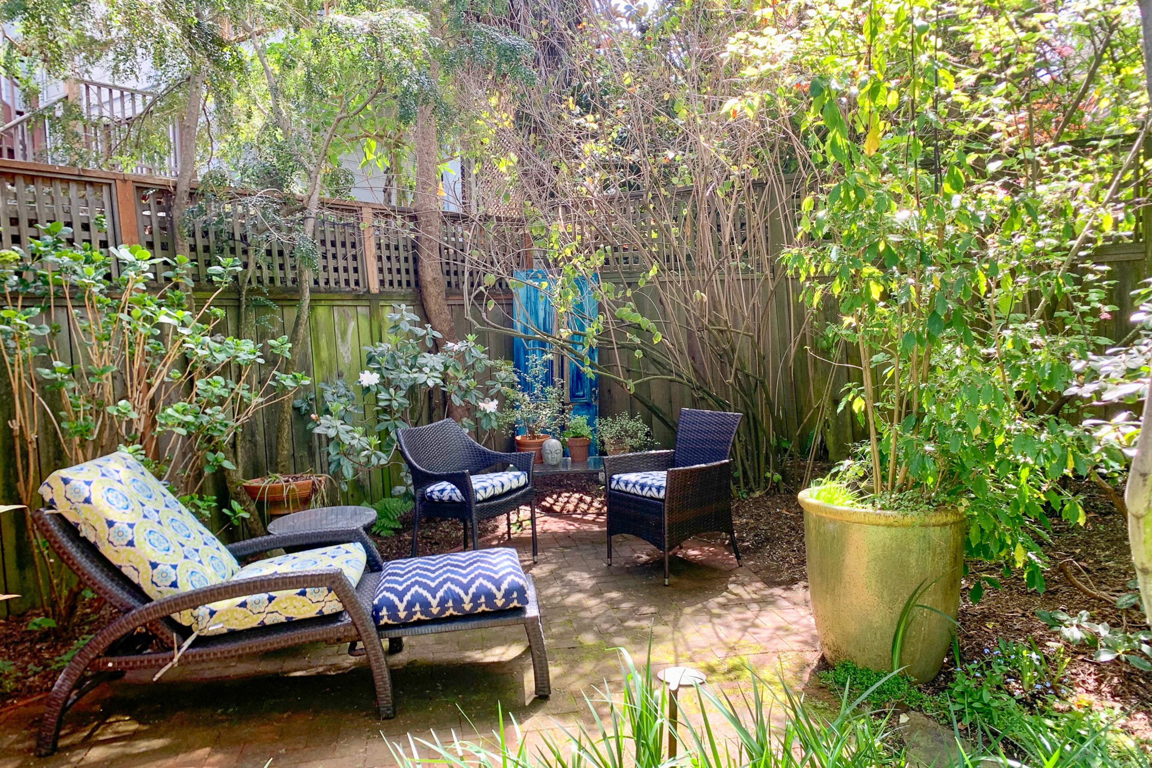 A cozy garden seating area with wicker chairs and cushions is surrounded by lush green plants and a wooden fence under dappled sunlight.
