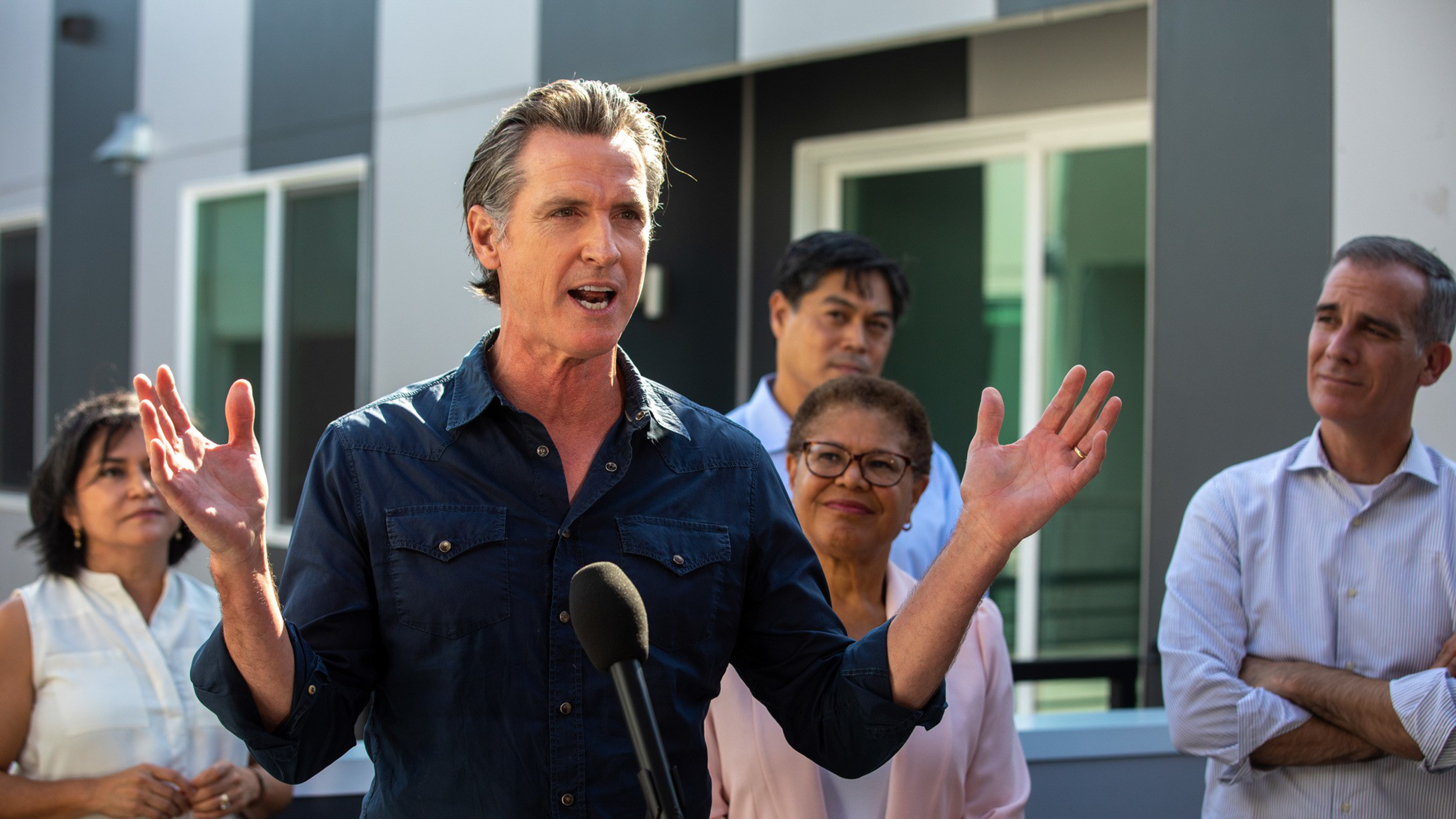 A man in a dark shirt speaks passionately at a microphone while four people behind him listen attentively outside a modern building.