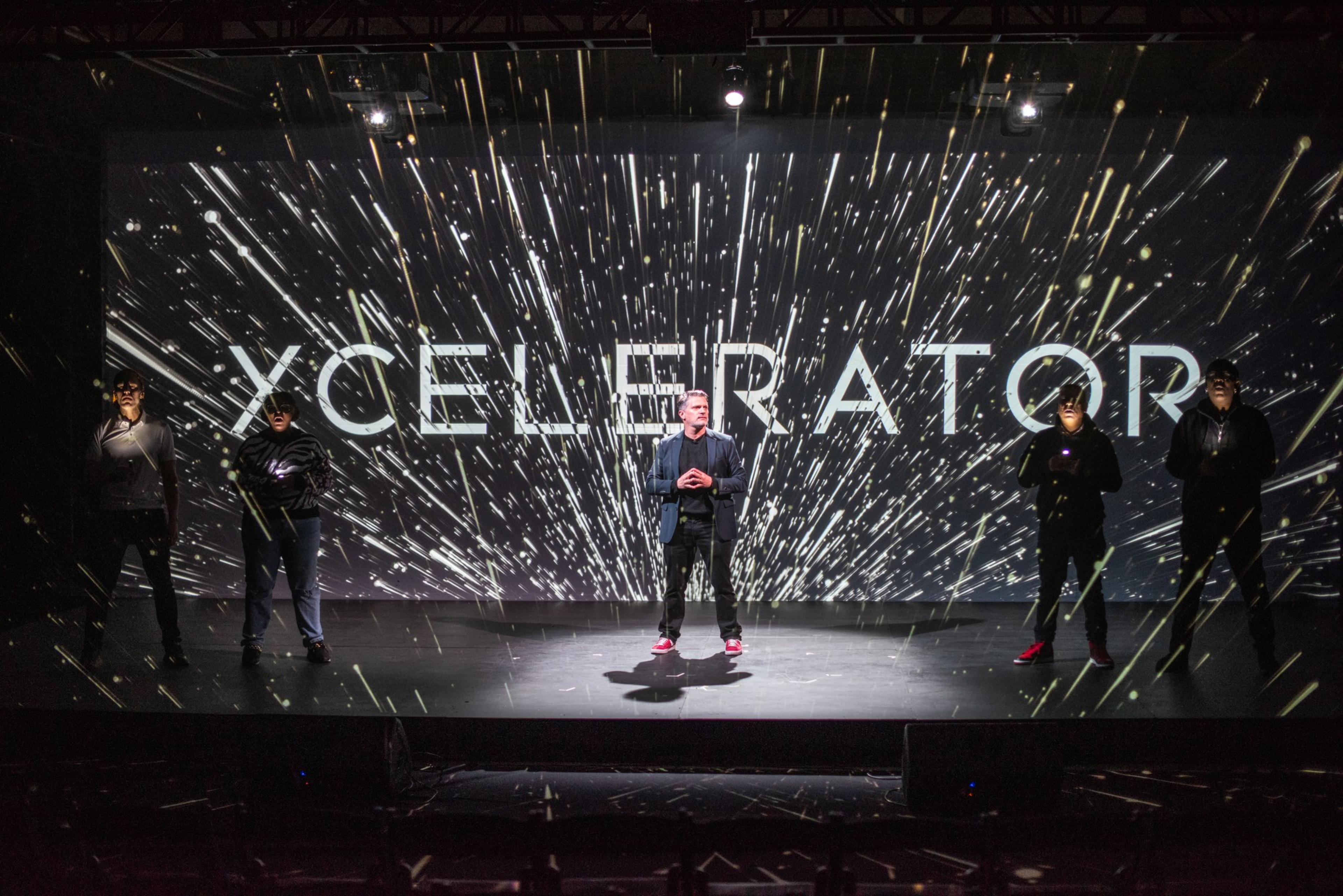 Five people stand on a stage with dynamic light effects and the word &quot;XCELERATOR&quot; displayed prominently behind them.