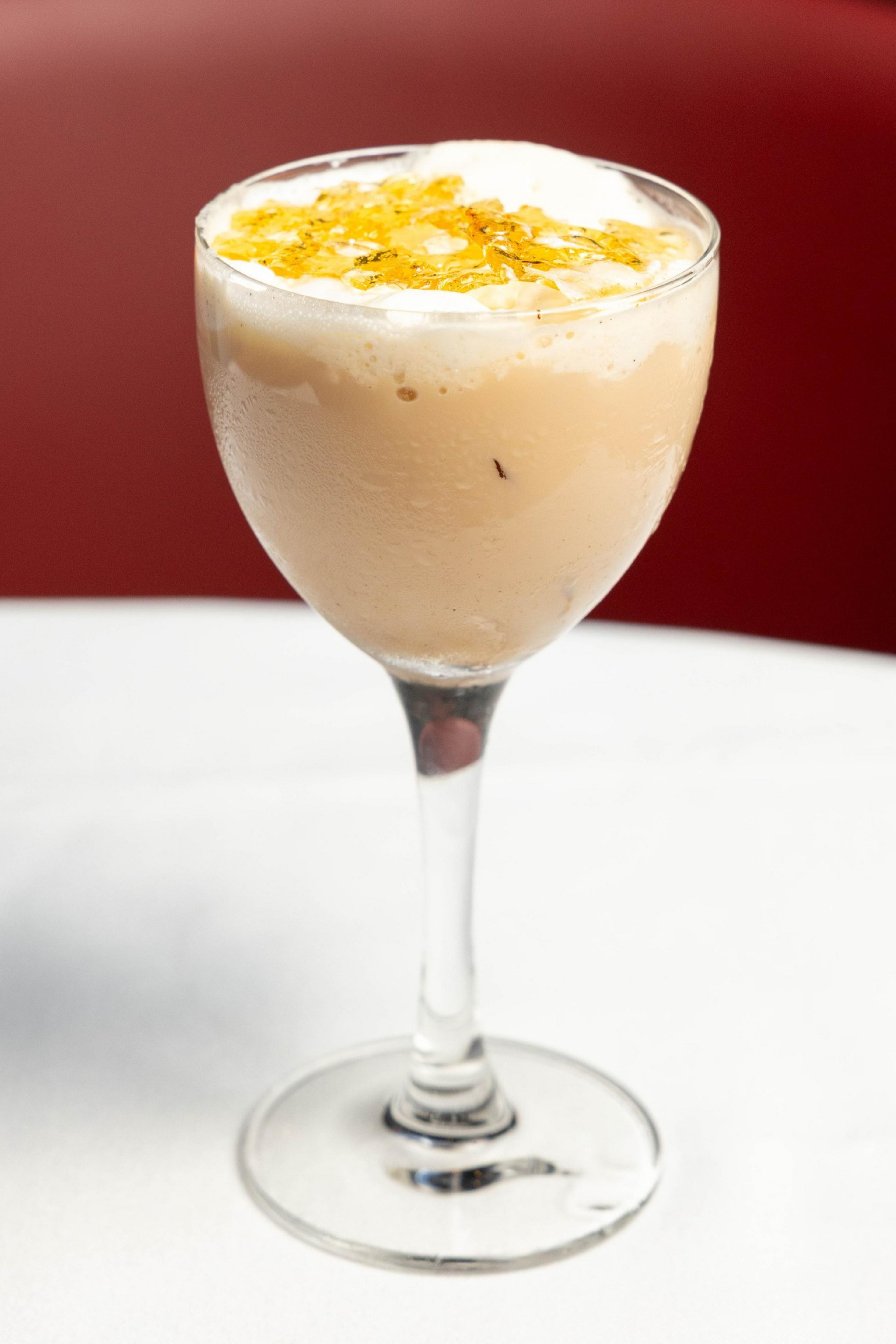 A creamy cocktail in a stemmed glass is topped with golden shards, likely caramel, on a smooth, frothy surface, placed on a white table.