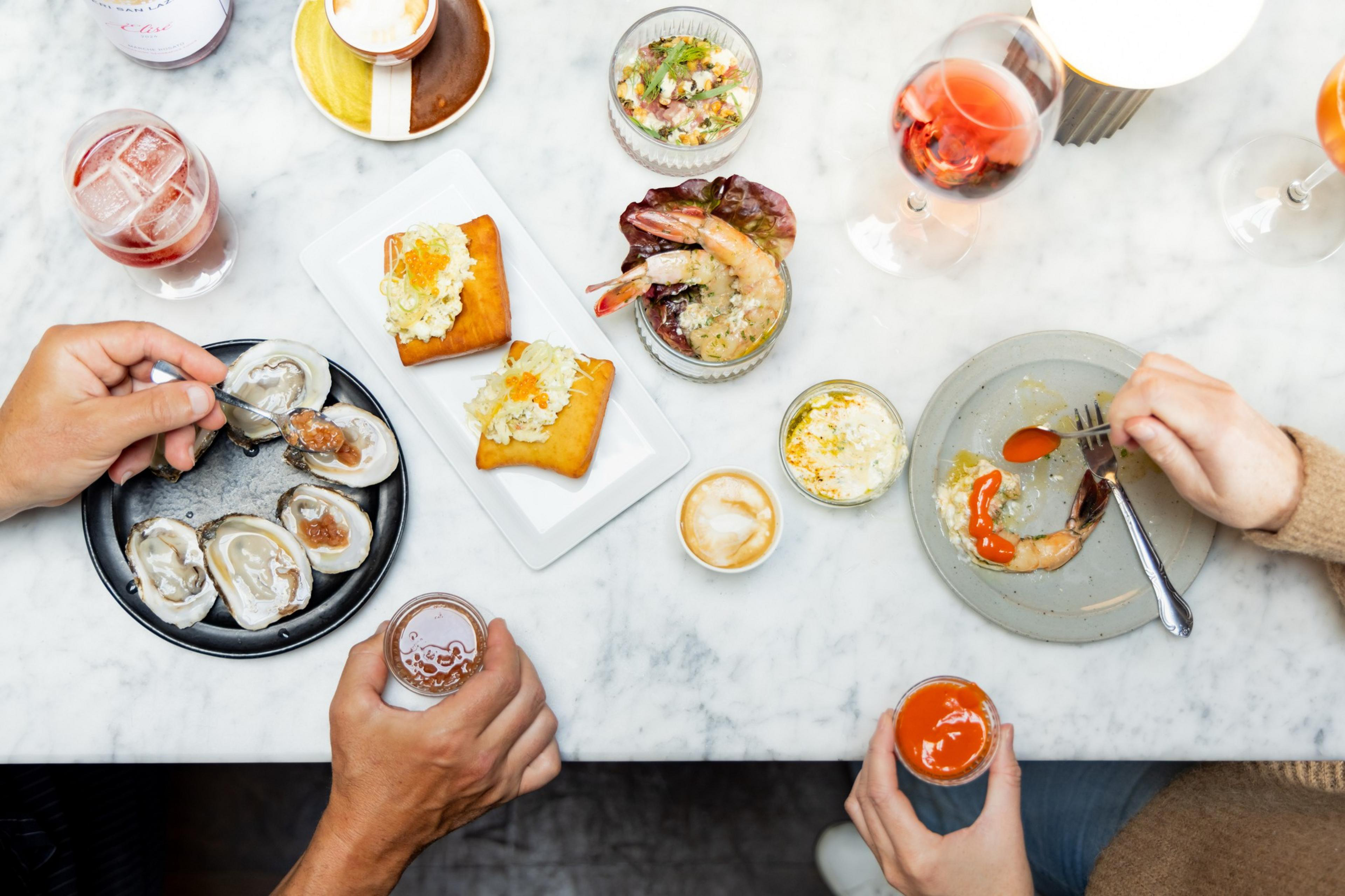 A marble table with oysters, shrimp, tart with topping, vibrant cocktails, and dipping sauces. Hands are preparing to eat, creating an elegant dining scene.