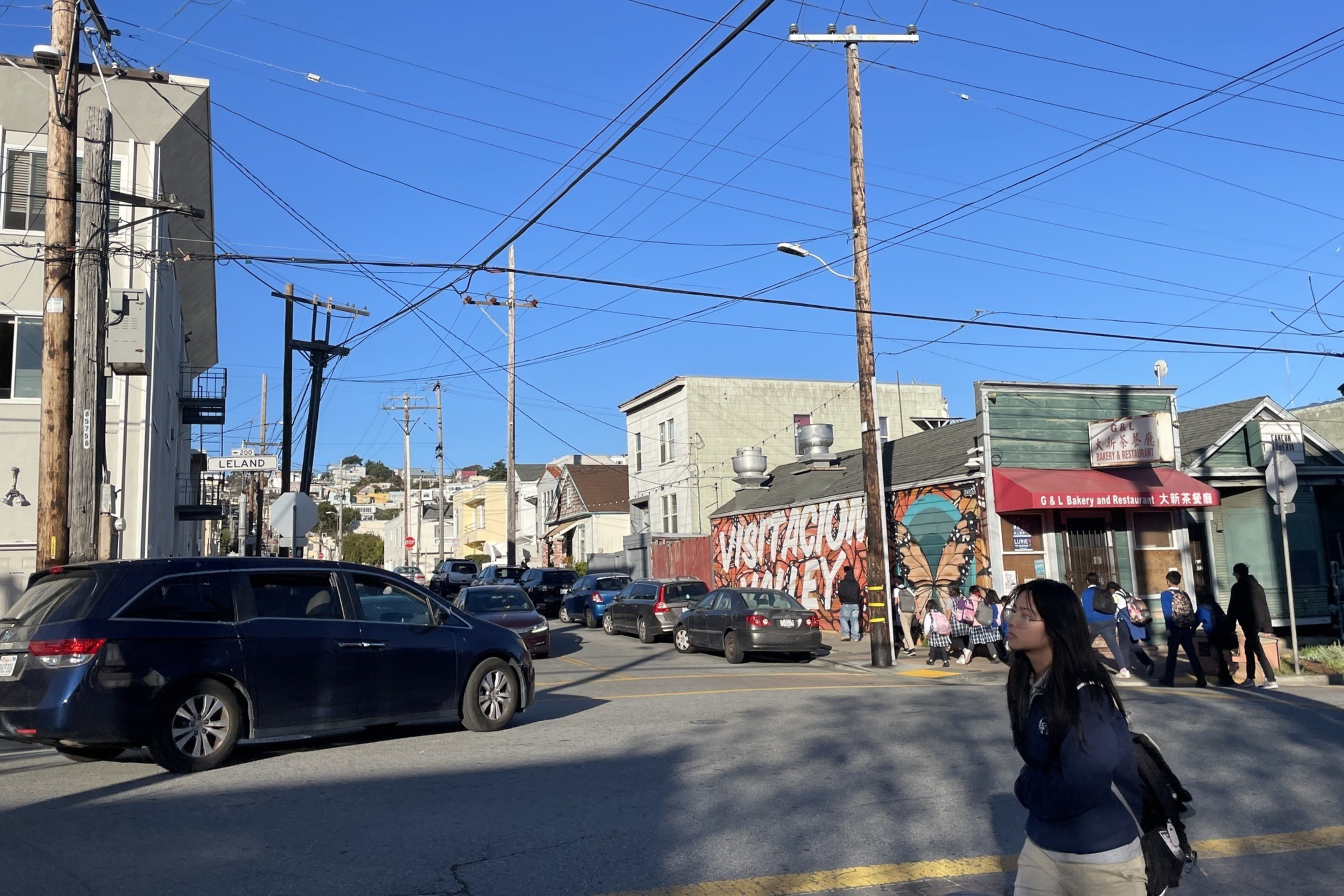 A busy street intersection with a van and cars. A bakery is nearby, and a mural reads "Visitacion Valley." A person with a backpack walks in the foreground.