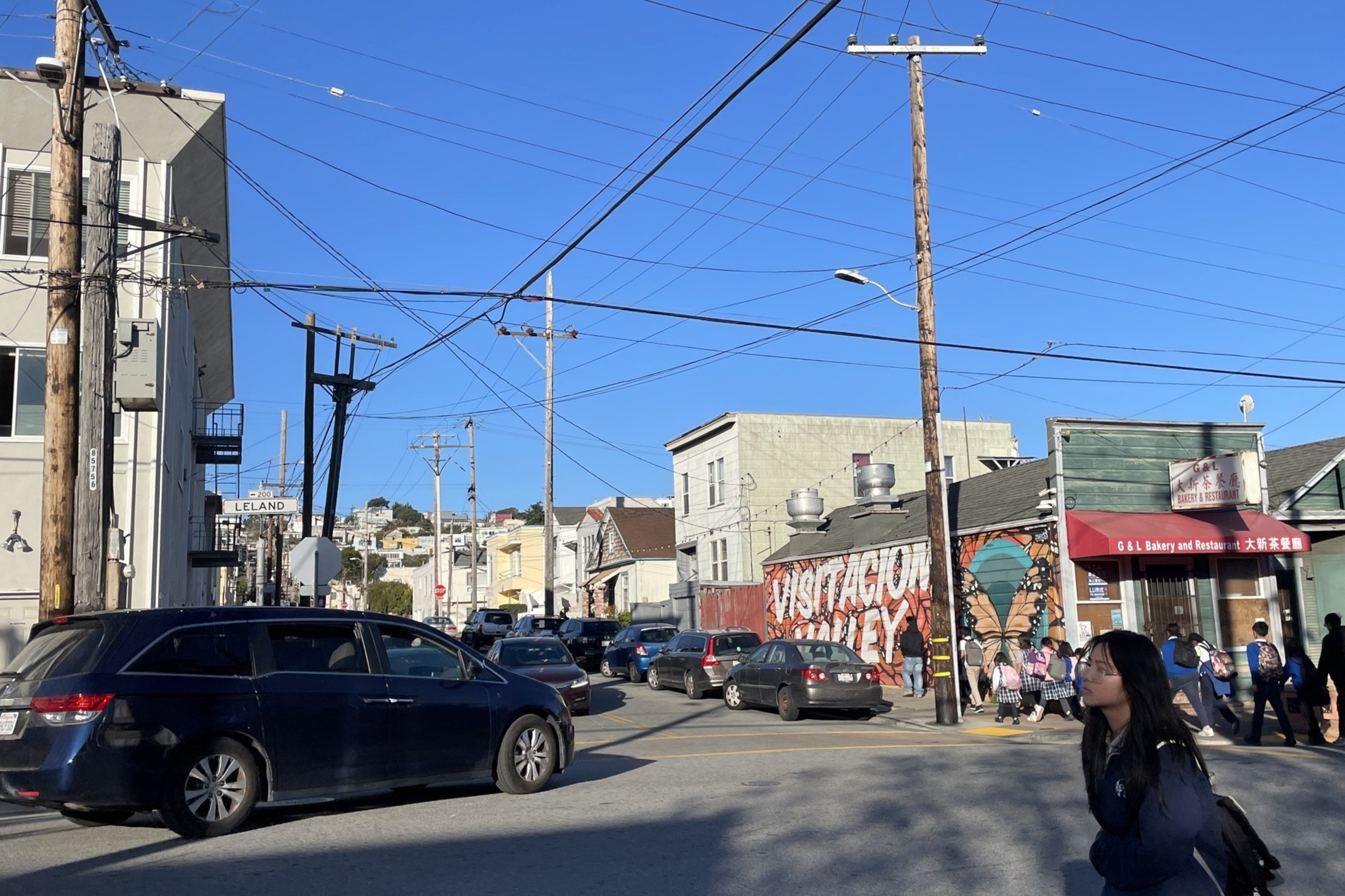 A busy street intersection with a van and cars. A bakery is nearby, and a mural reads "Visitacion Valley." A person with a backpack walks in the foreground.
