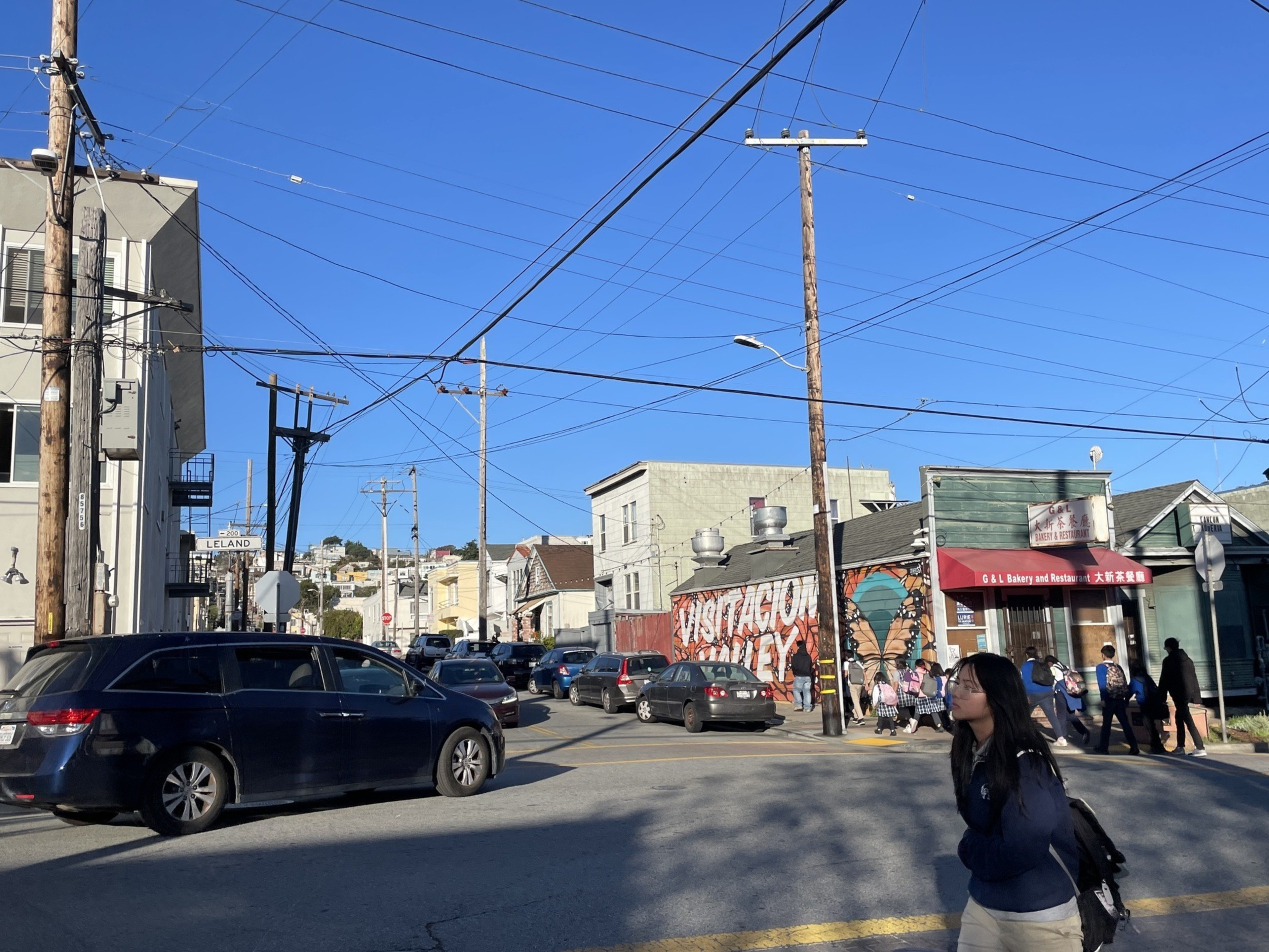 A busy street intersection with a van and cars. A bakery is nearby, and a mural reads "Visitacion Valley." A person with a backpack walks in the foreground.