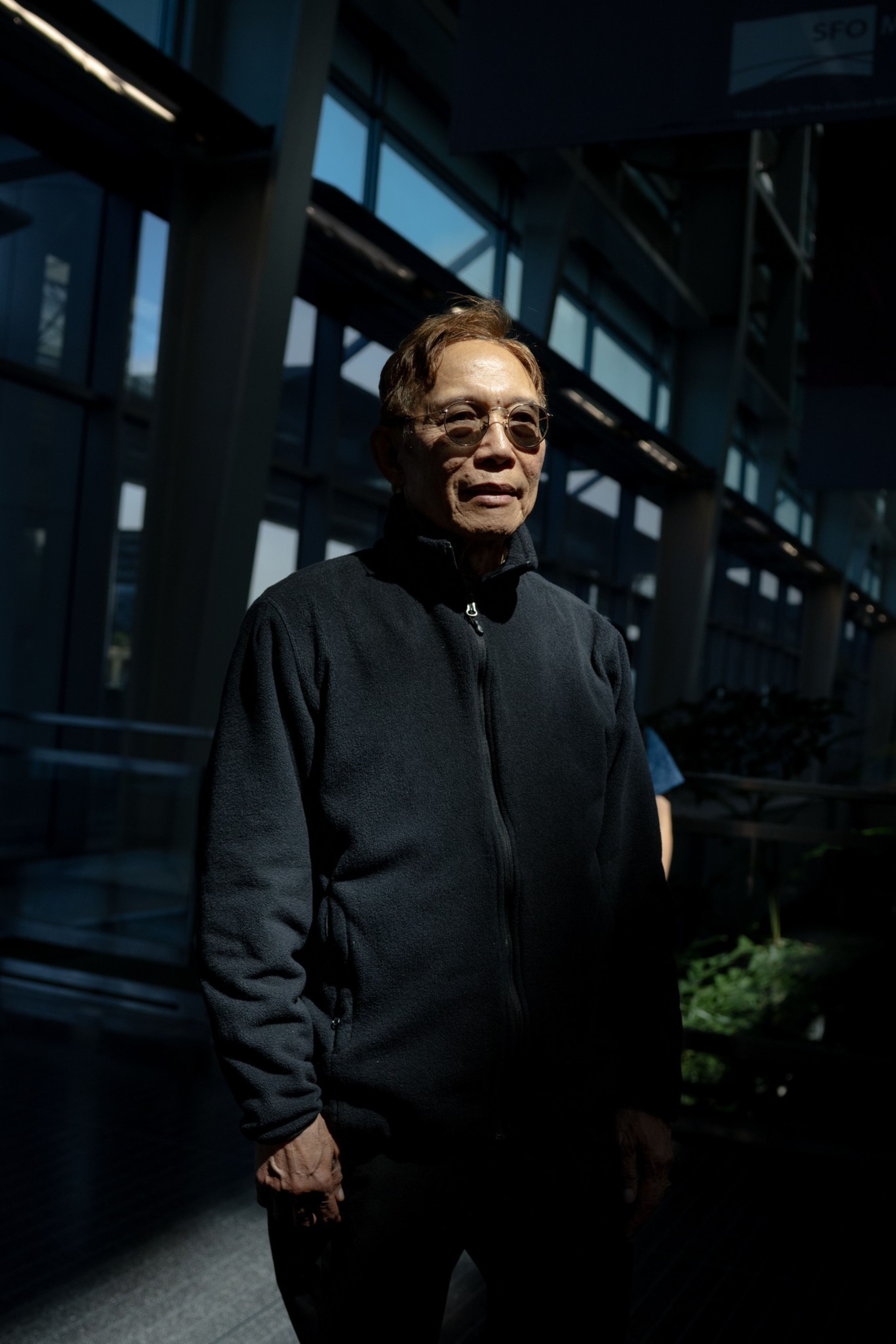 A man wearing glasses and a dark zip-up jacket stands in a dimly lit modern building with large windows and steel beams.