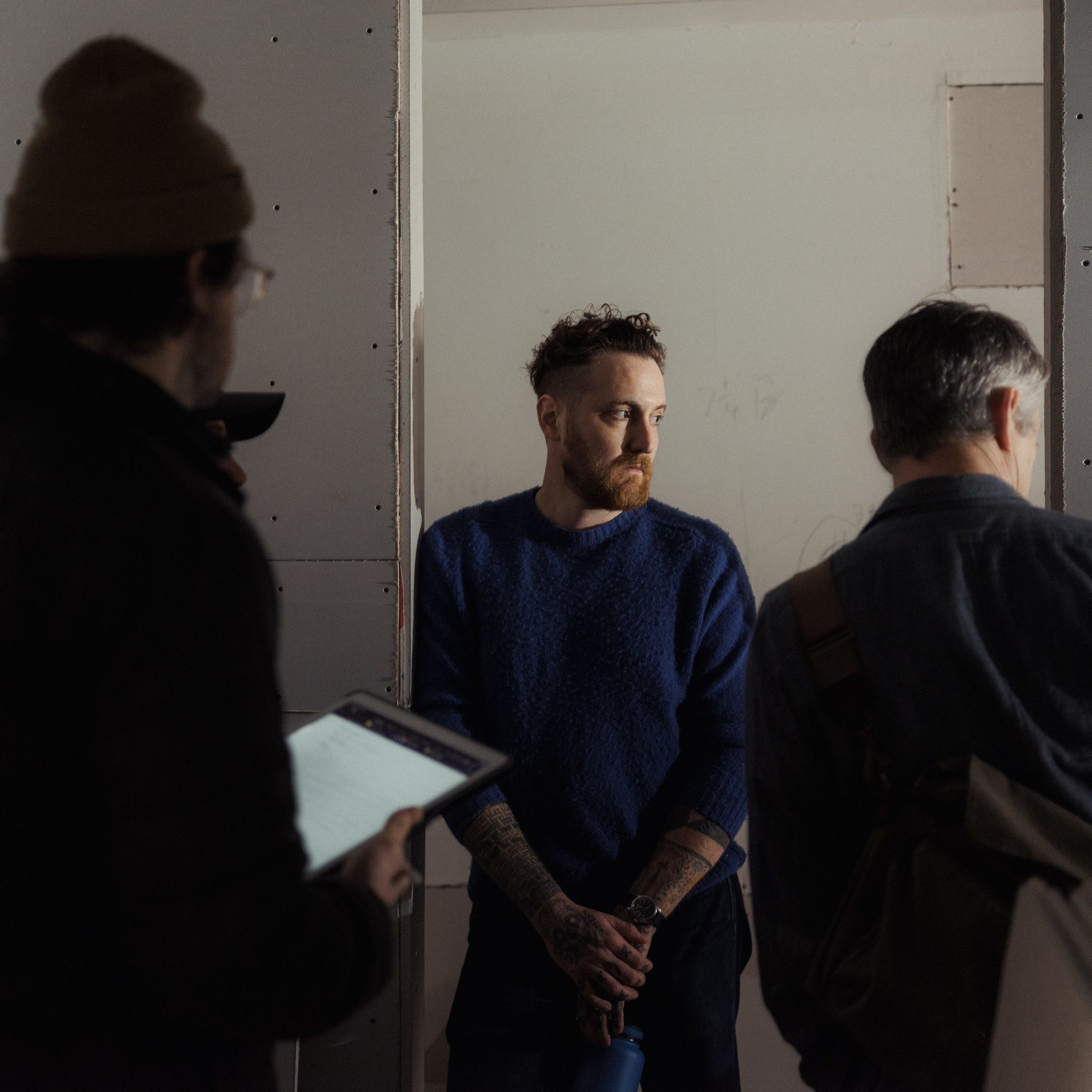 A man in a blue sweater, with tattoos, stands against a wall holding a bottle. Two other people are partially visible, one with a clipboard.