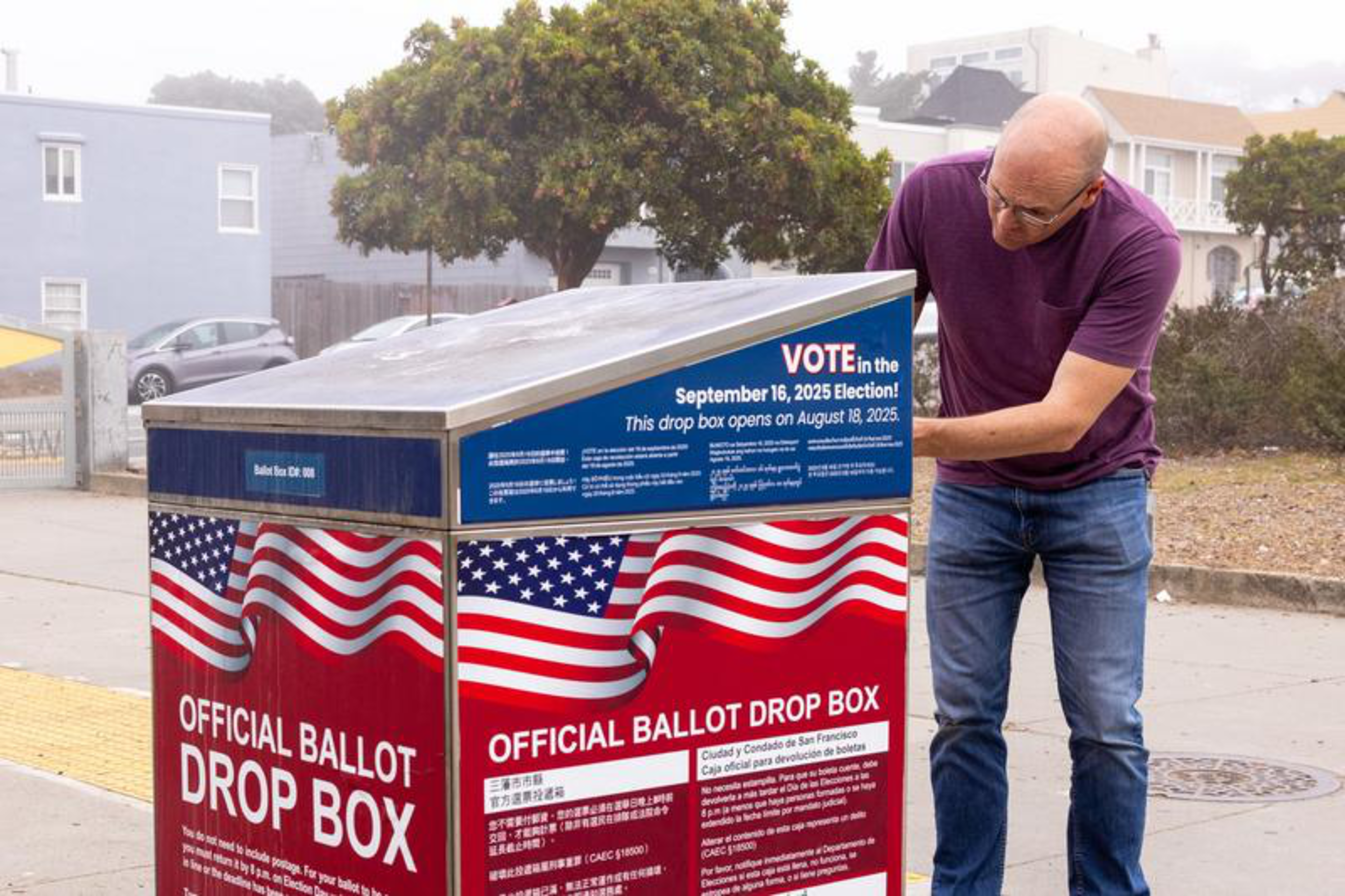 A man in a purple shirt and jeans is dropping a ballot into an official red, white, and blue ballot drop box for the September 16, 2025 election.