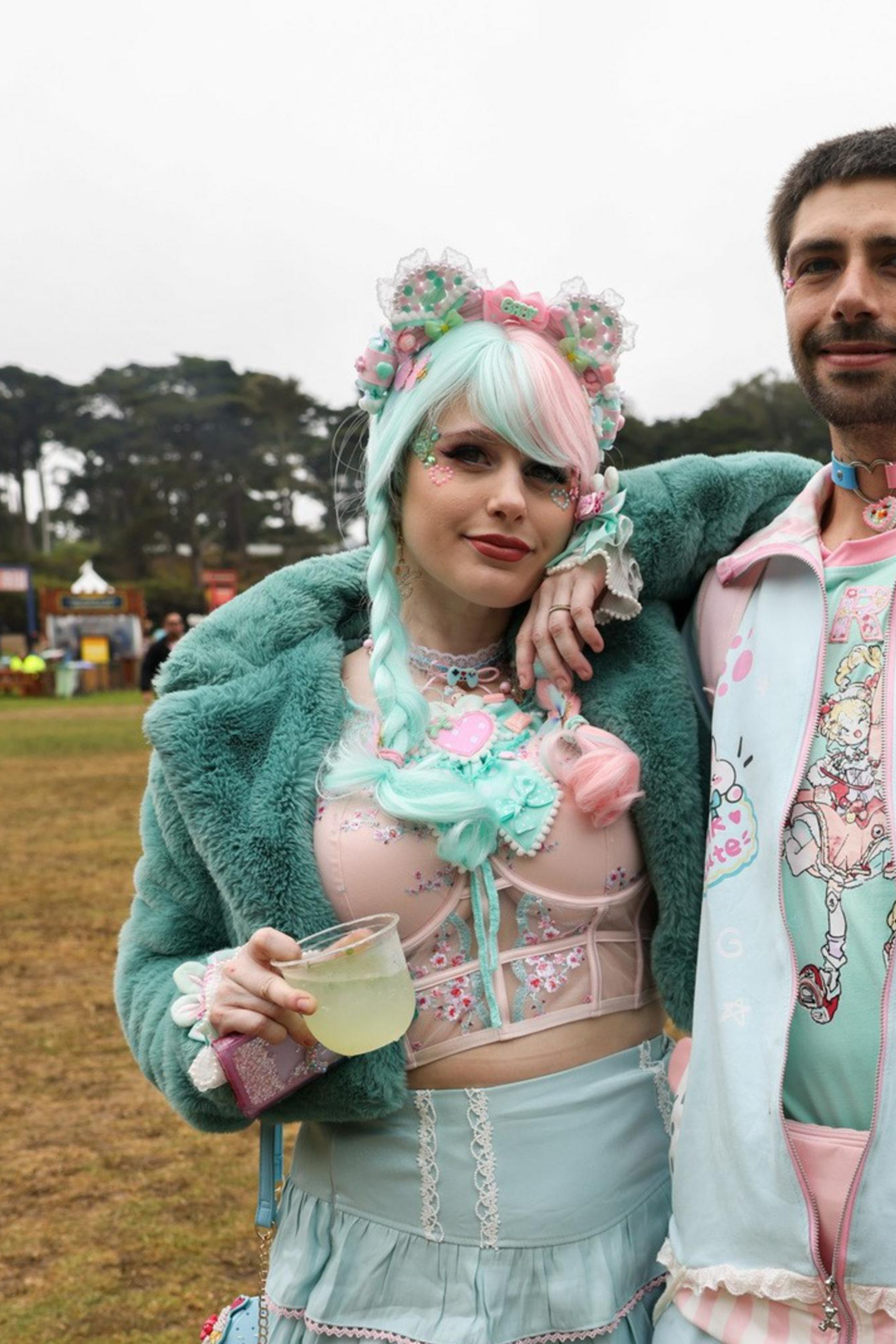 A colorful couple poses at an outdoor event. The woman wears a pastel goth-style outfit with turquoise and pink accents, while the man sports matching pastel attire.