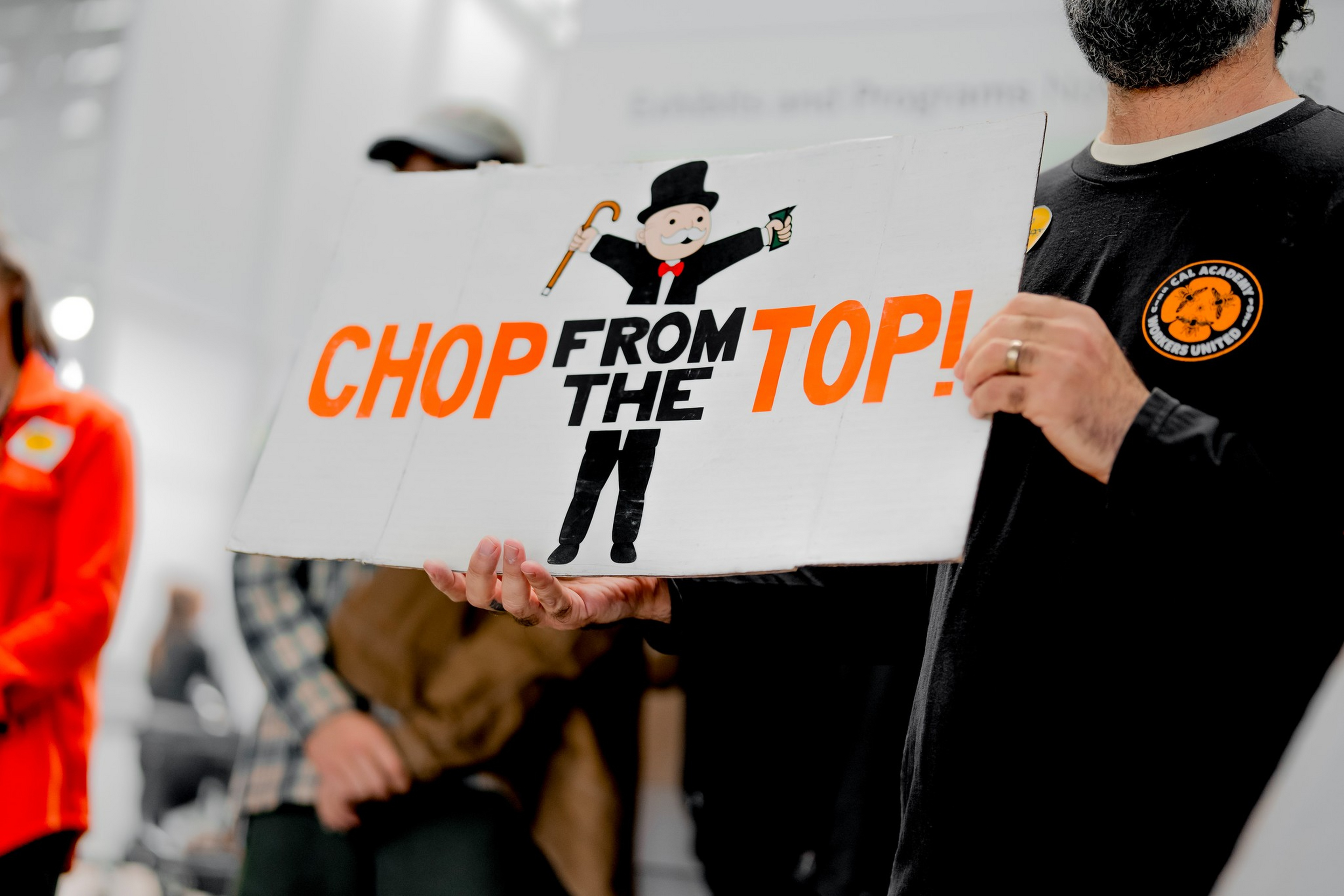 A person holds a sign reading “CHOP FROM THE TOP!” with an illustration of a Monopoly man holding a cane and money.