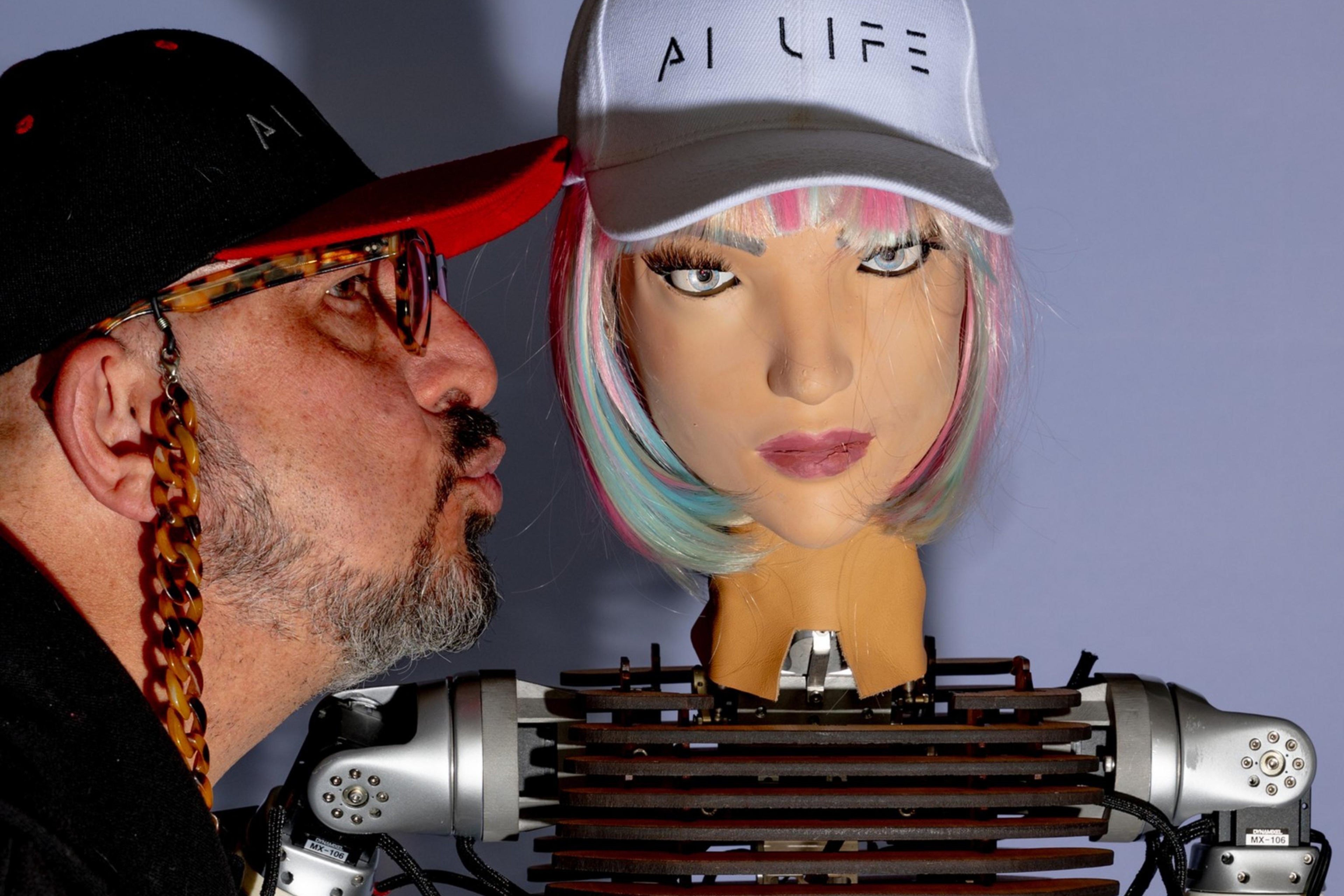 A man wearing glasses and a cap leans in to kiss a humanoid robot with colorful hair, both wearing caps labeled "AI LIFE." The robot has a visible mechanical body.