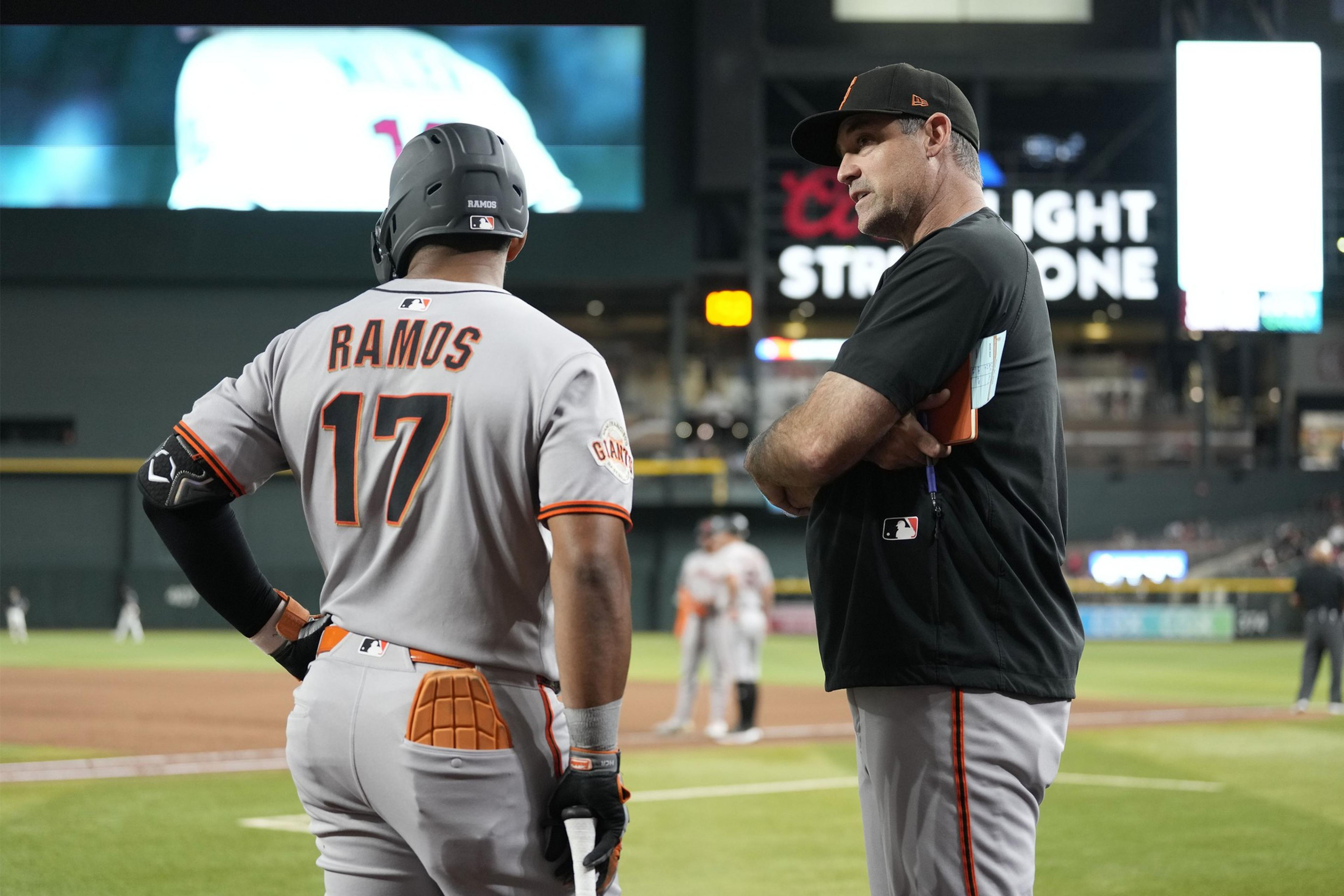 A baseball player wearing a gray Giants uniform with "Ramos 17" talks to a coach in black, standing near the field during a nighttime game.