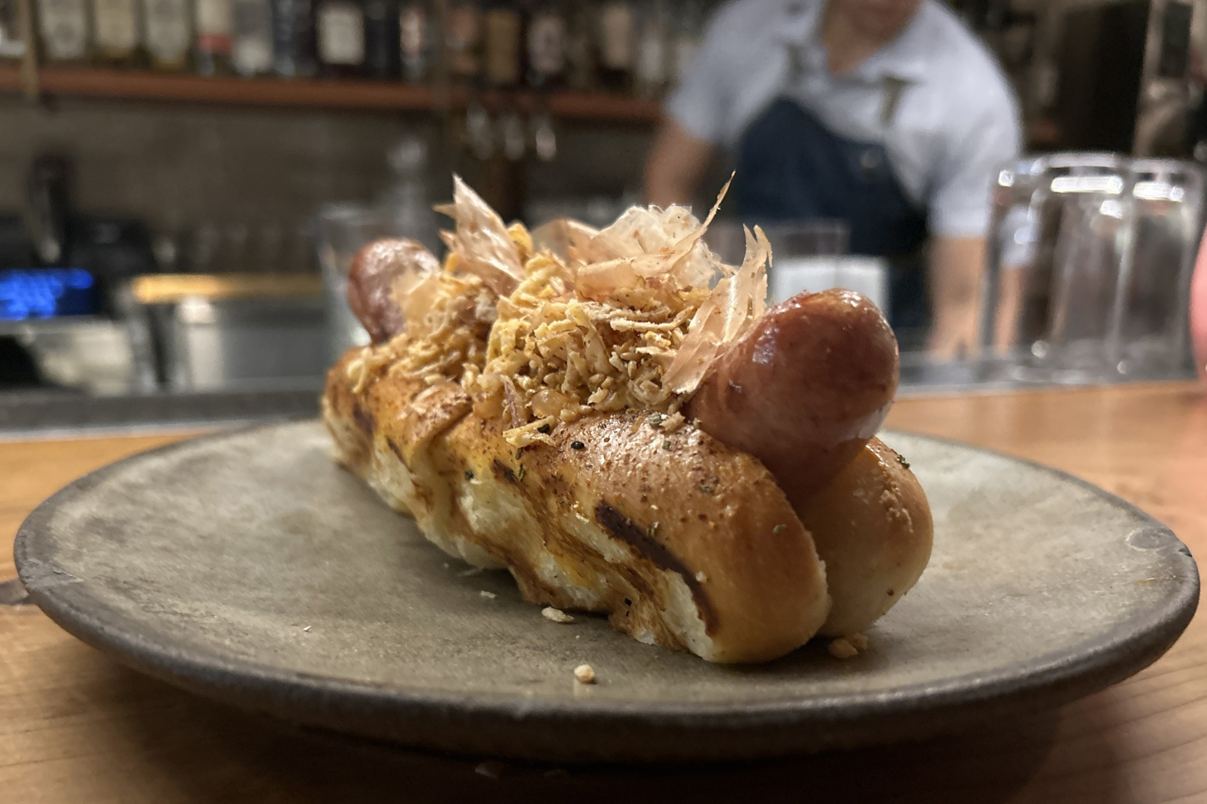 A low angle view of a hotdog topped with kimchi, bonito flakes, toasted sesame, fried shallots and rosemary aioli on a togarashi bun.