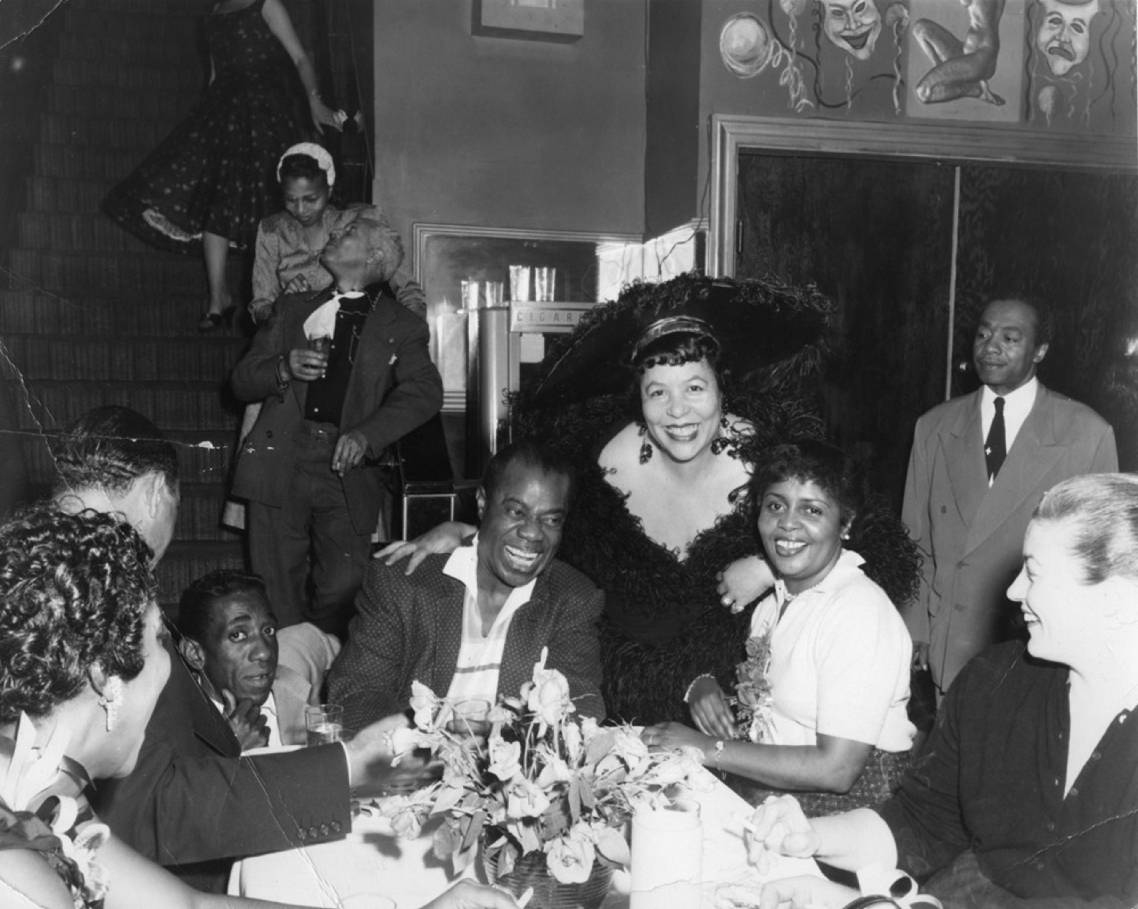 A smiling woman in a large feathered hat stands behind a table where people laugh and chat, while others mingle and descend stairs in the background.
