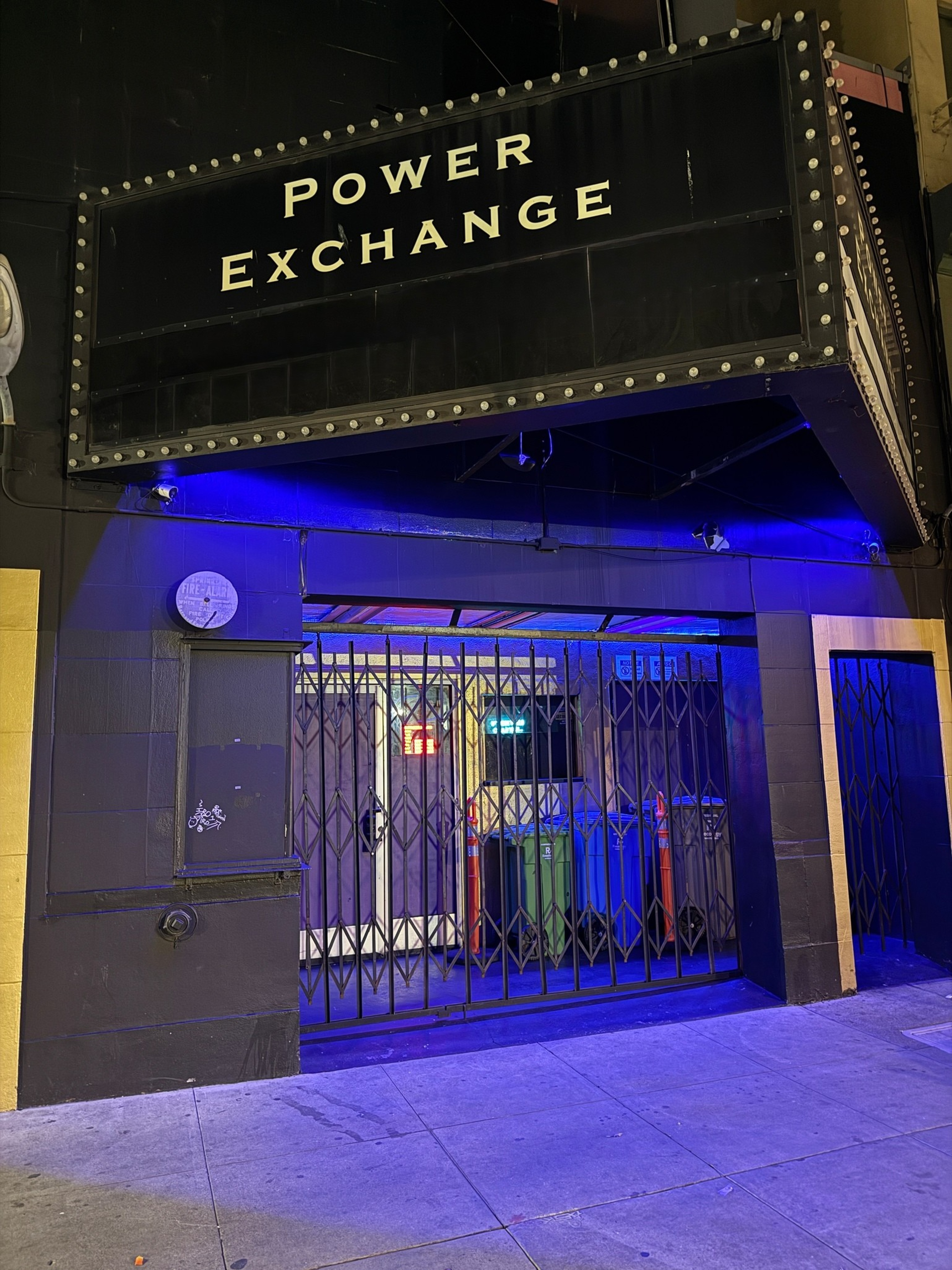 A closed venue with a black sign reading “POWER EXCHANGE” above a gated entrance lit by blue light, showing some trash bins and caution cones inside.