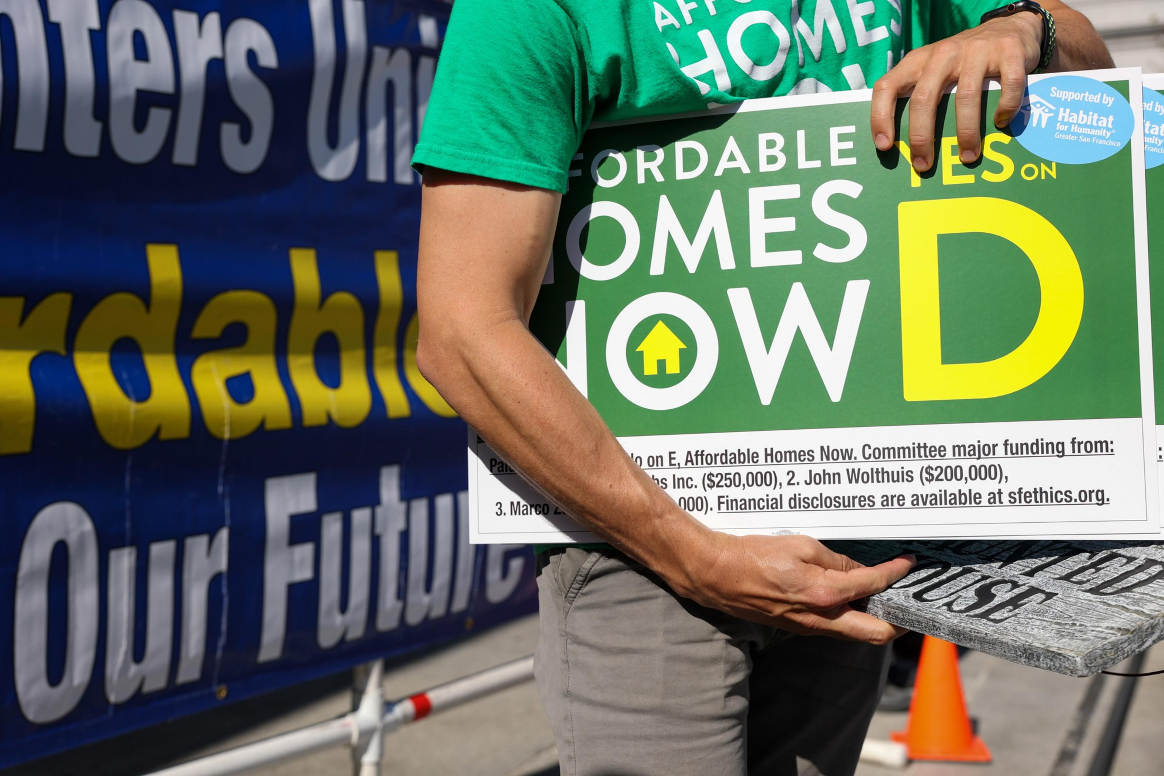 A person in a green "Affordable Homes" shirt holds a sign advocating for affordable homes on E. Yes on D, with funding details at the bottom.