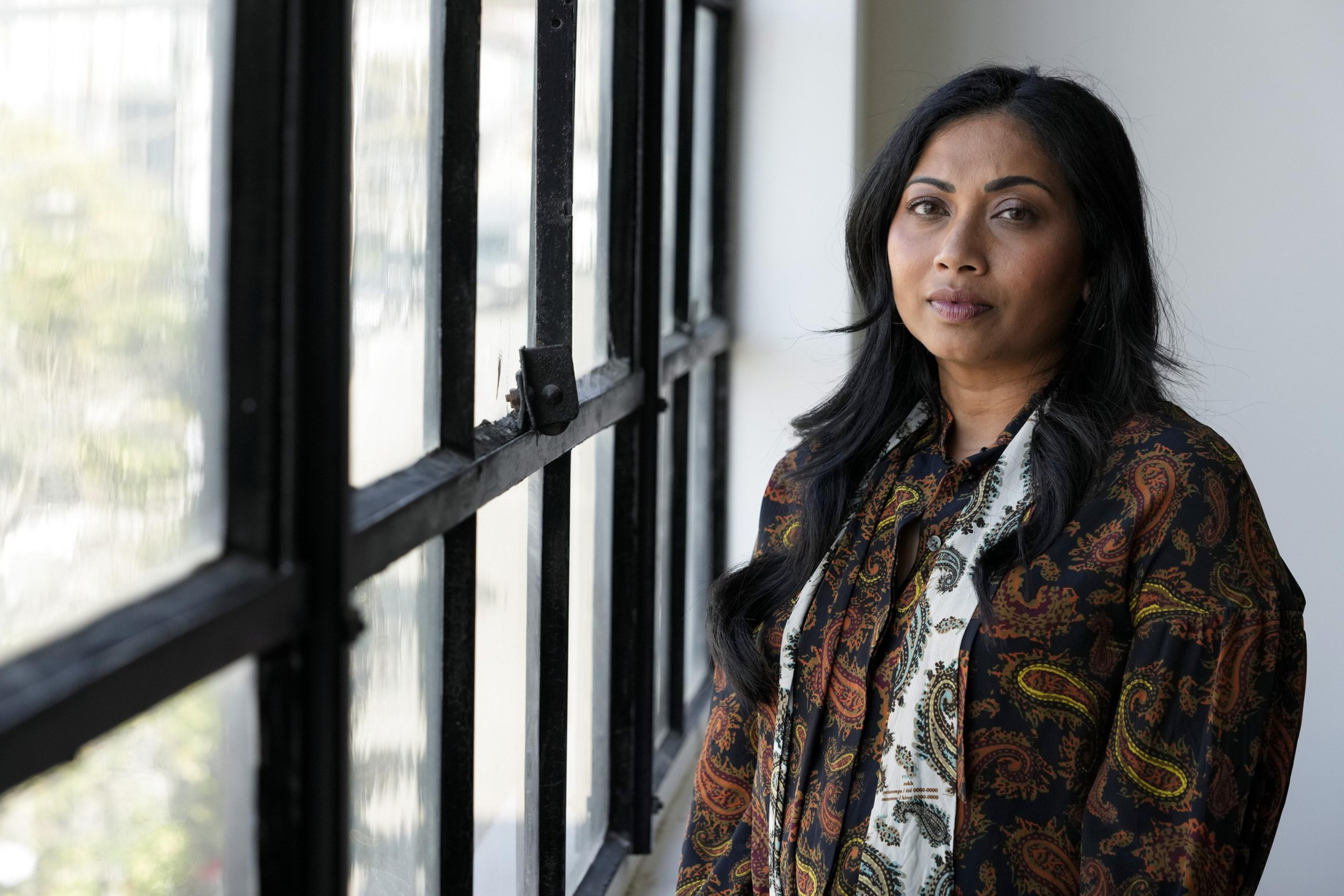 A person with long black hair stands next to a window.