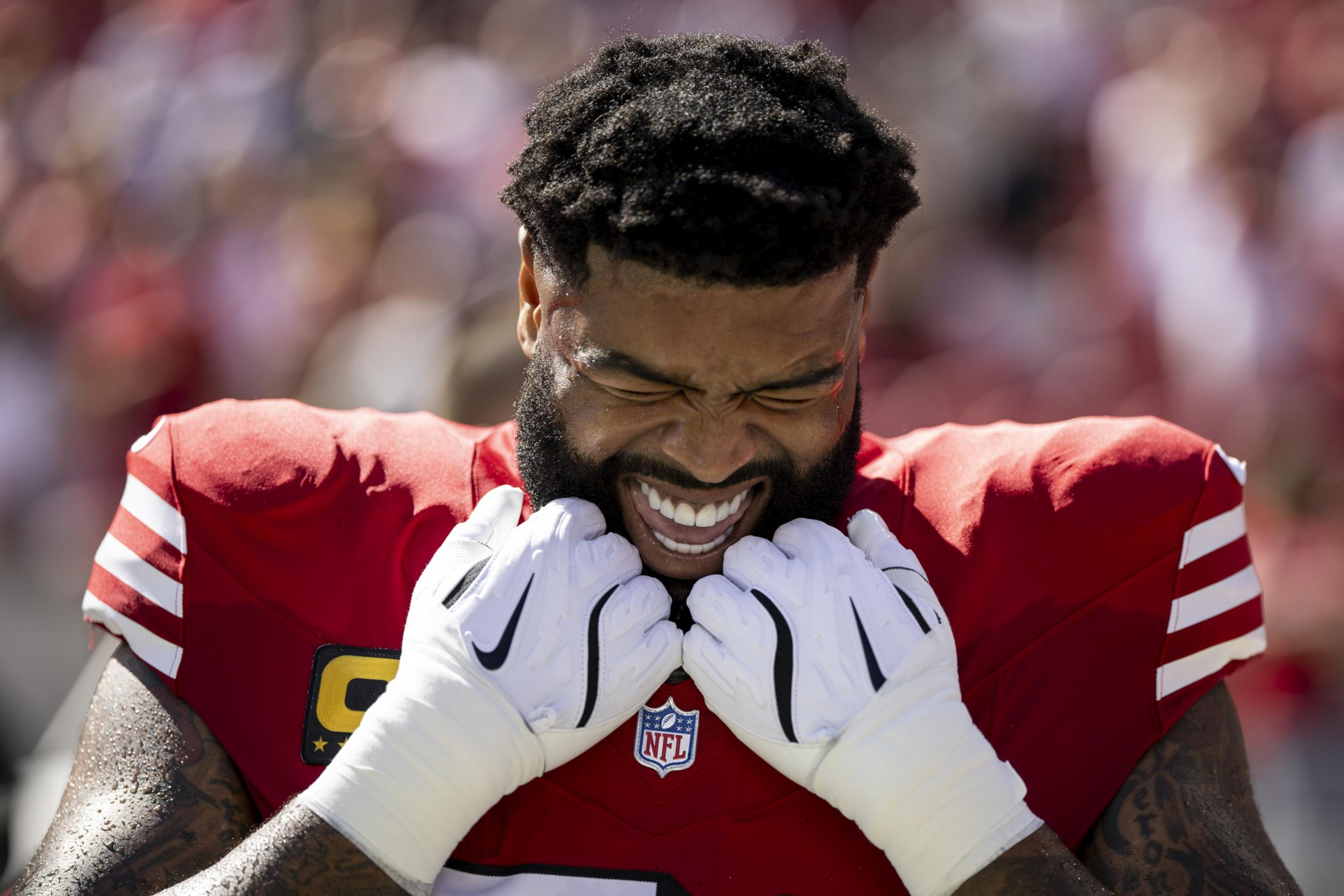A football player in a red jersey and white gloves stands with eyes closed, teeth clenched, and fists near his face, seeming determined before a game.