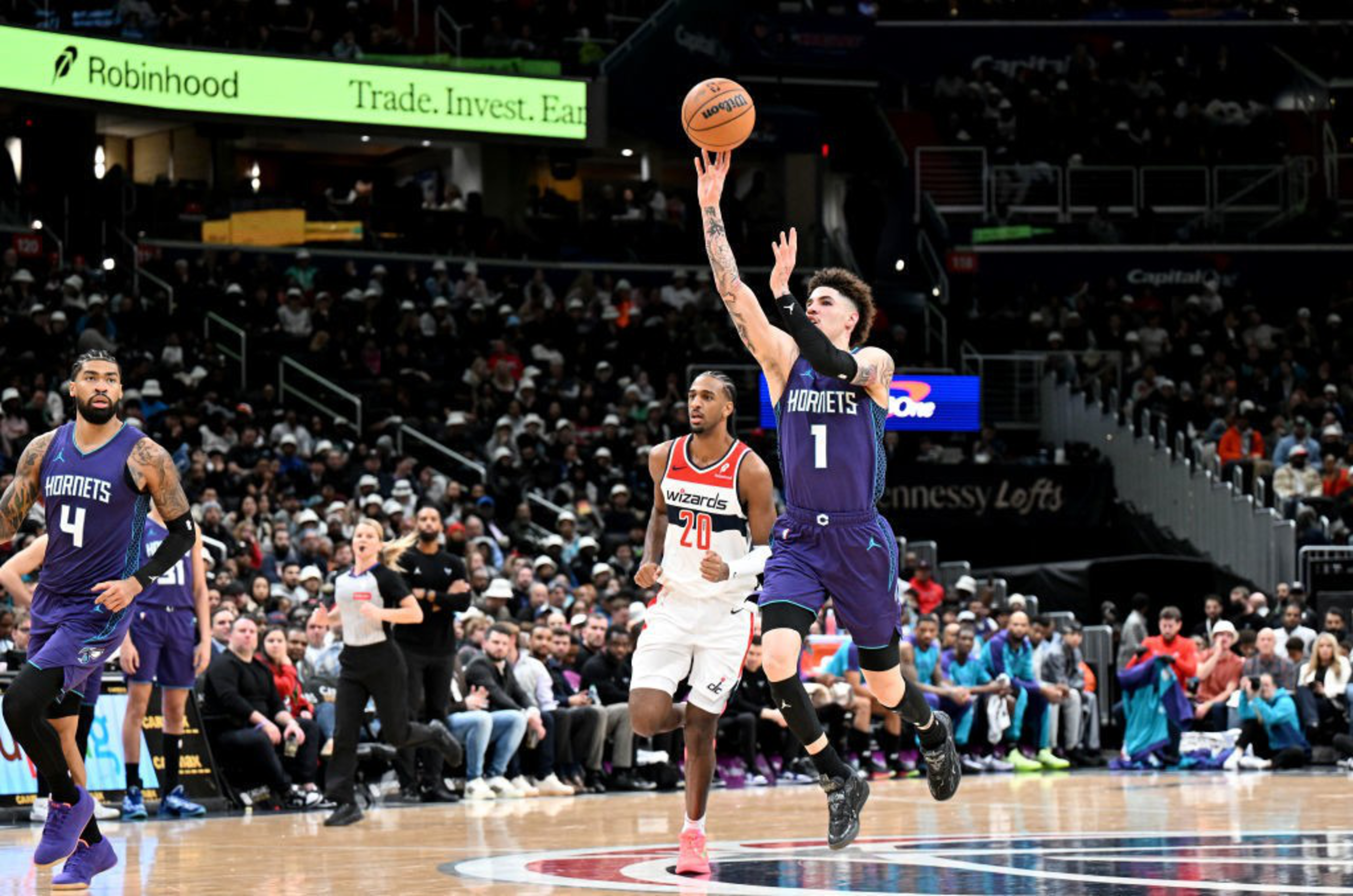 A basketball player in a purple Hornets jersey is leaping to pass the ball during a game. An opposing player in a white Wizards jersey is close behind.