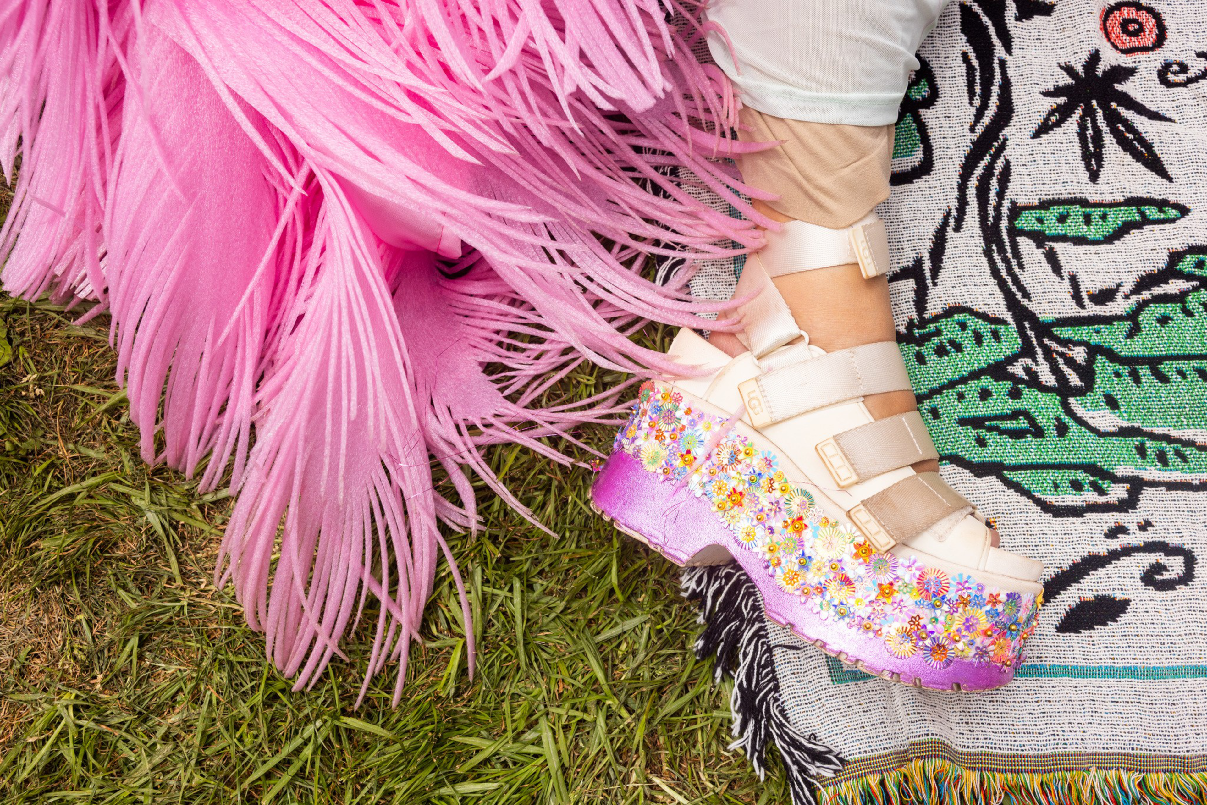A foot wearing a white platform sandal with floral decorations rests on a colorful patterned rug next to long pink fringed fabric on grass.