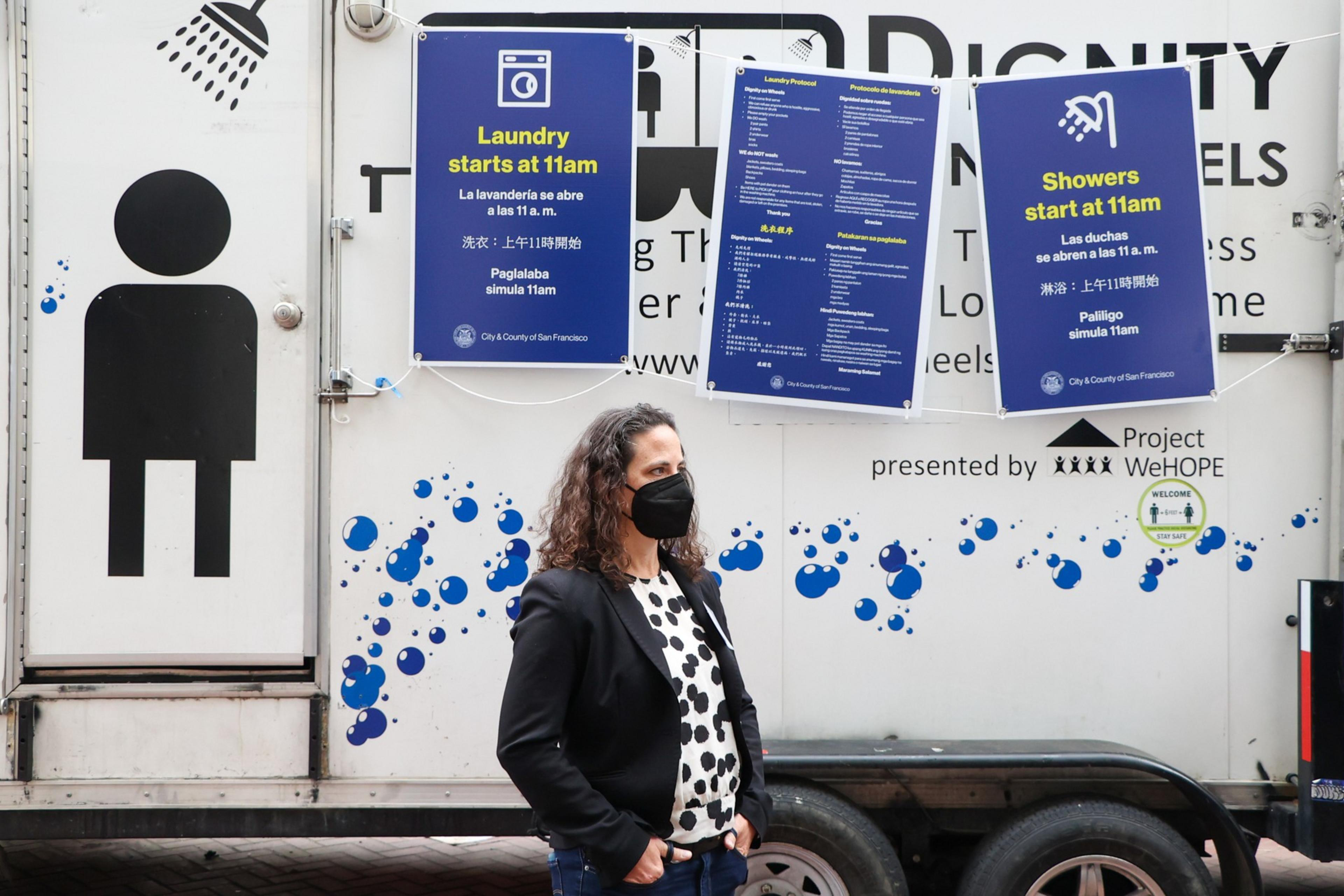 A woman wearing a black mask and polka dot shirt stands in front of a mobile shower and laundry truck with signs stating laundry and showers start at 11am.