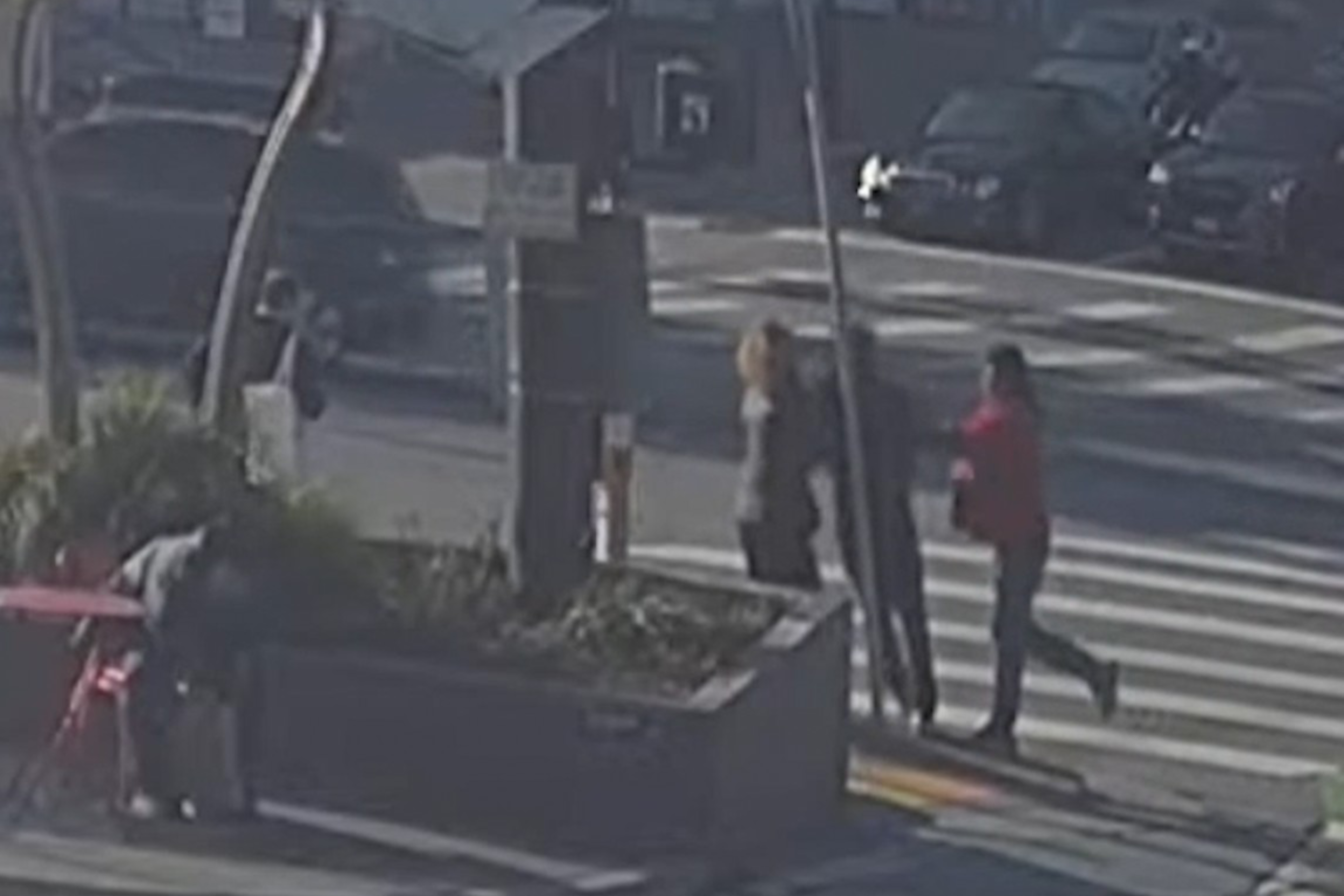 Three people are crossing a street at a crosswalk. A fourth person is hunched over near a red table. Cars and other pedestrians are visible in the background.