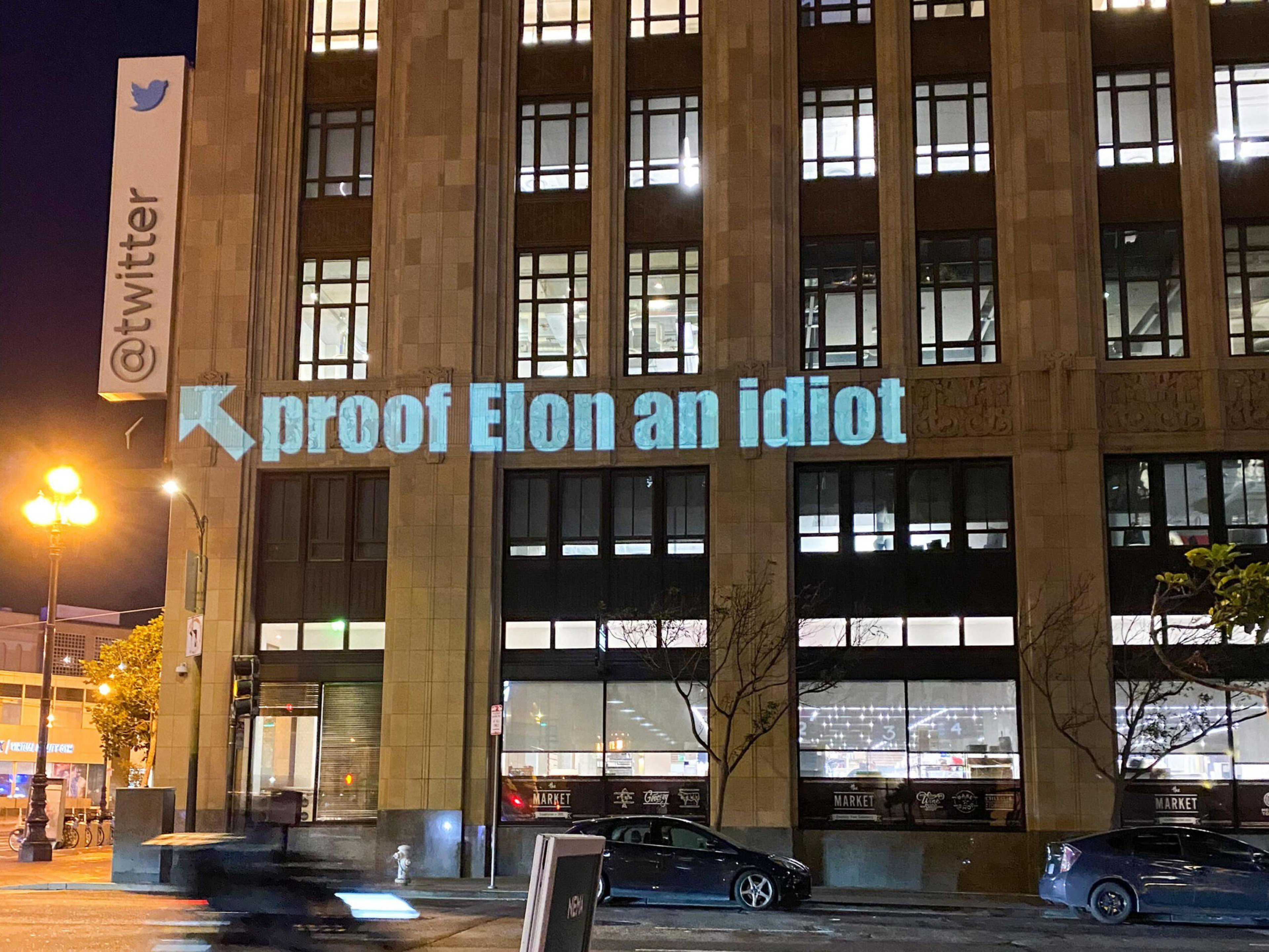 A projection on a building near a Twitter sign reads "proof Elon an idiot" with an arrow pointing to the left, against a nighttime city backdrop.