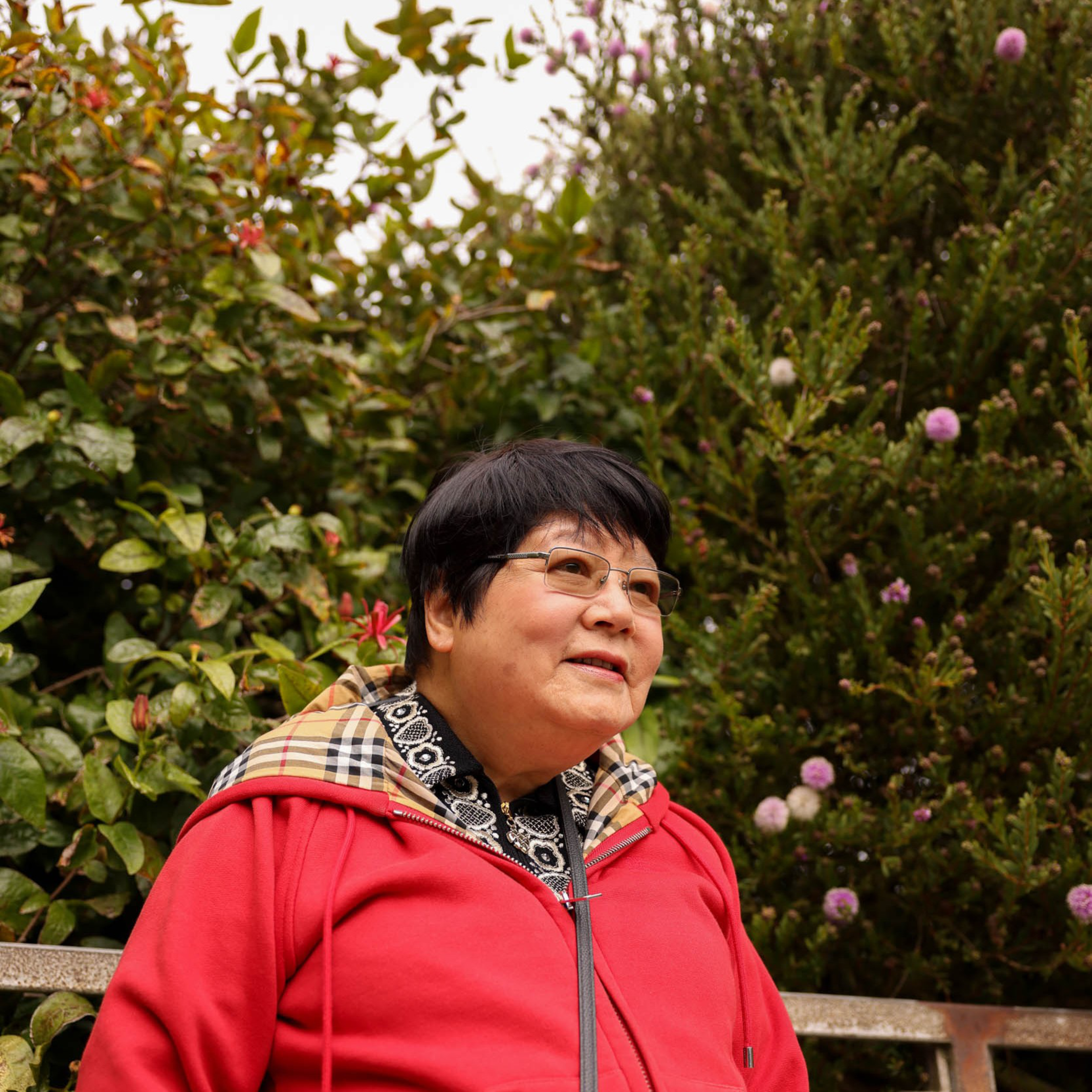 An older woman with short black hair and glasses wears a red jacket and patterned scarf, sitting on a bench surrounded by green plants and purple flowers.