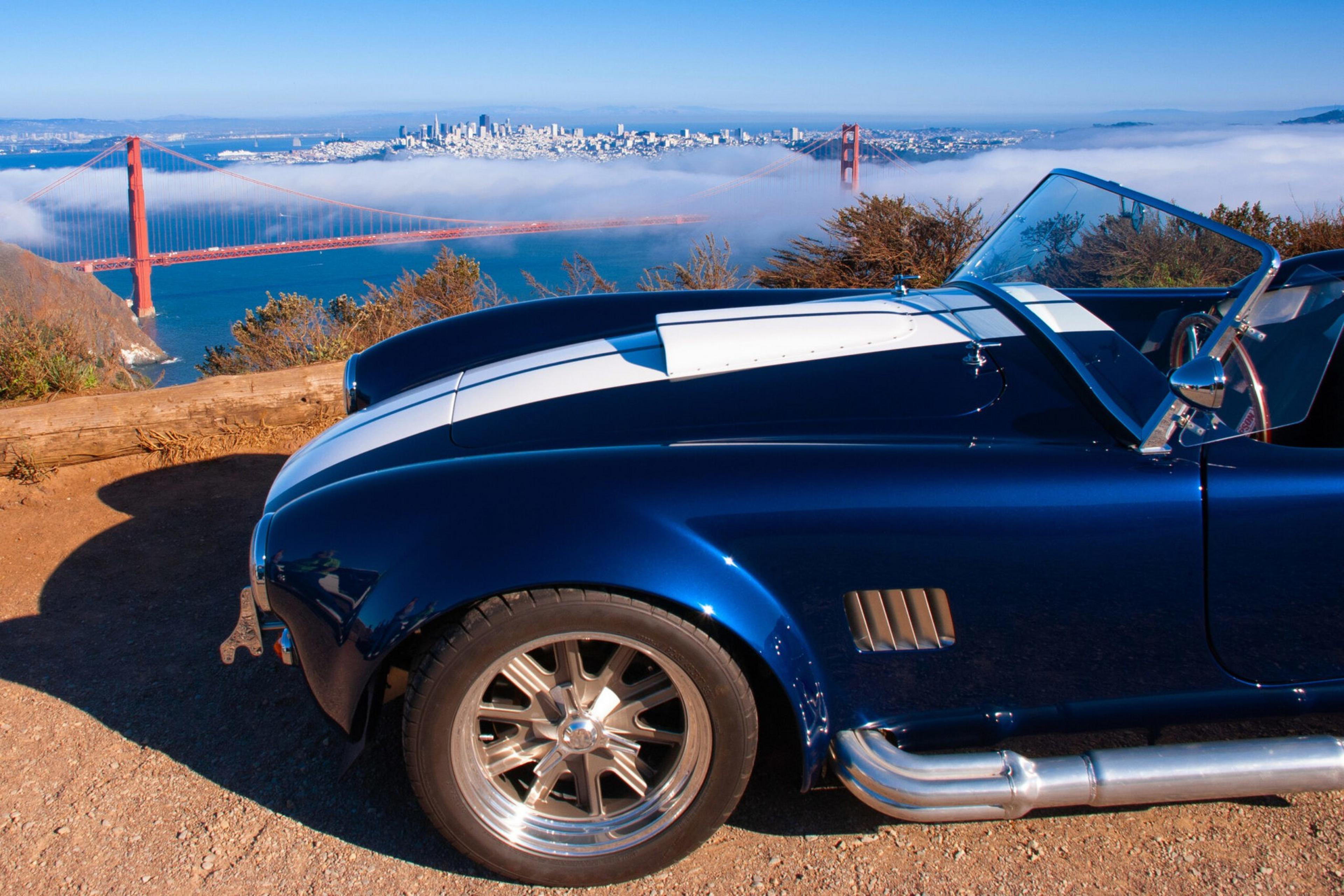A dark blue vintage sports car with white racing stripes is parked on a dirt overlook, with the Golden Gate Bridge and San Francisco skyline visible through fog in the background.