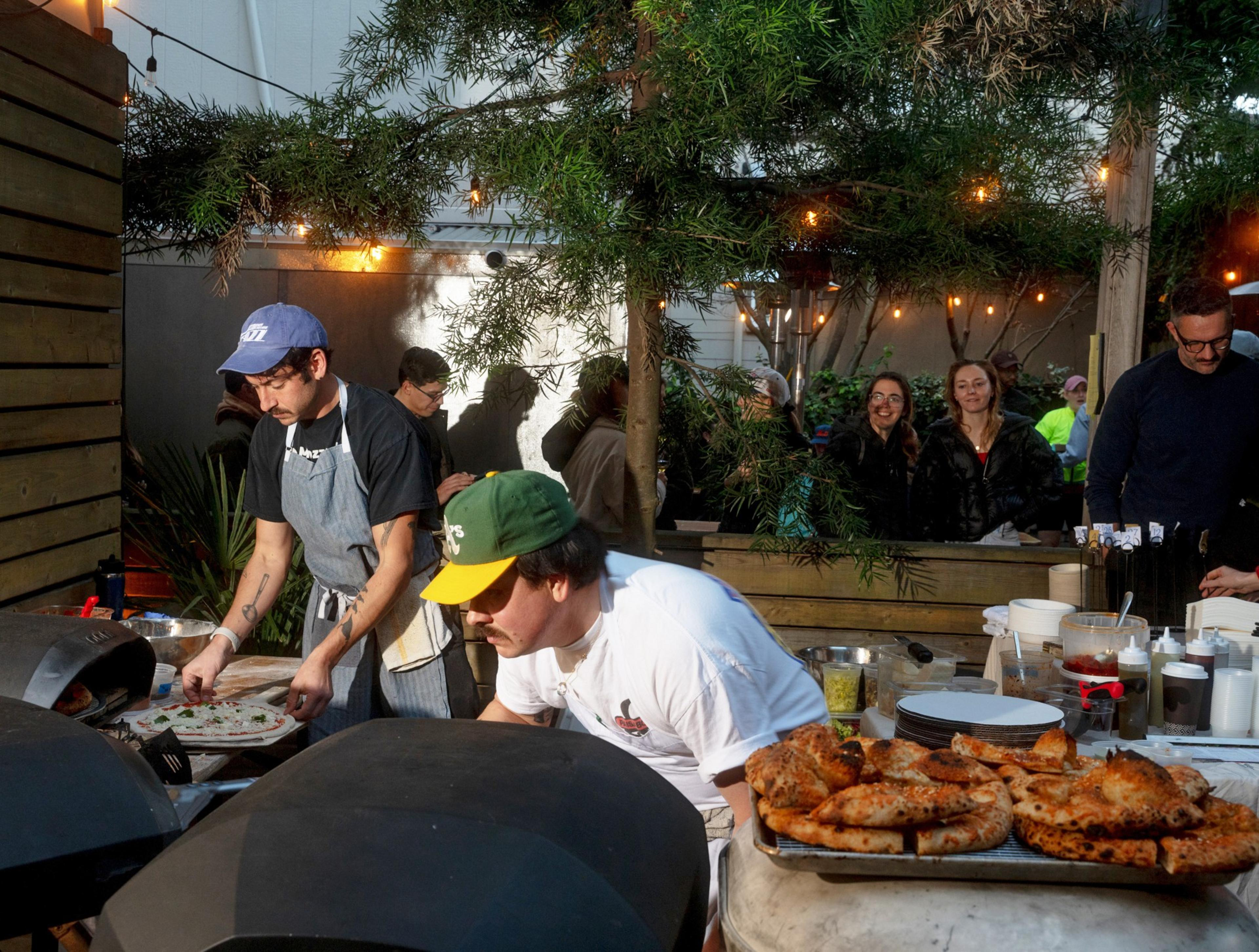 Two people make pizzas at an outdoor food stall, surrounded by plants and string lights. A table holds ingredients and baked pizzas, with onlookers nearby.